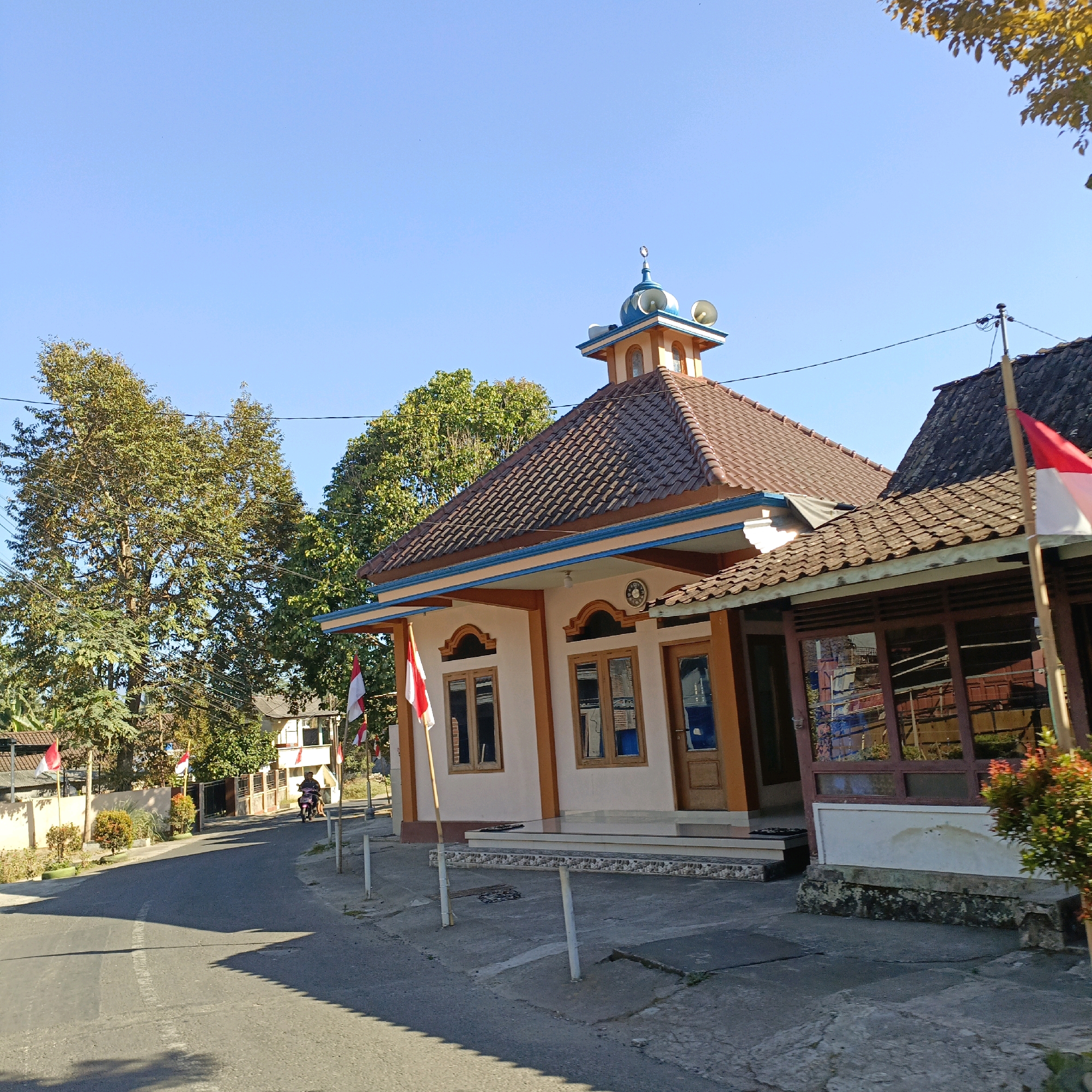 In Salatiga, Indonesia, on a clear, sunny day, a mosque with a distinctive blue-domed minaret stands prominently beside a paved road. The mosque features a brown tiled roof and cream-colored walls with orange trim. Indonesian flags, red and white, are visible on poles in front of the mosque. A motorcycle with a rider is moving away from the mosque on the road. Trees with green foliage line the street, and other buildings, some with traditional Indonesian architecture, are visible in the background. The scene captures a typical street view in a residential area of the city.