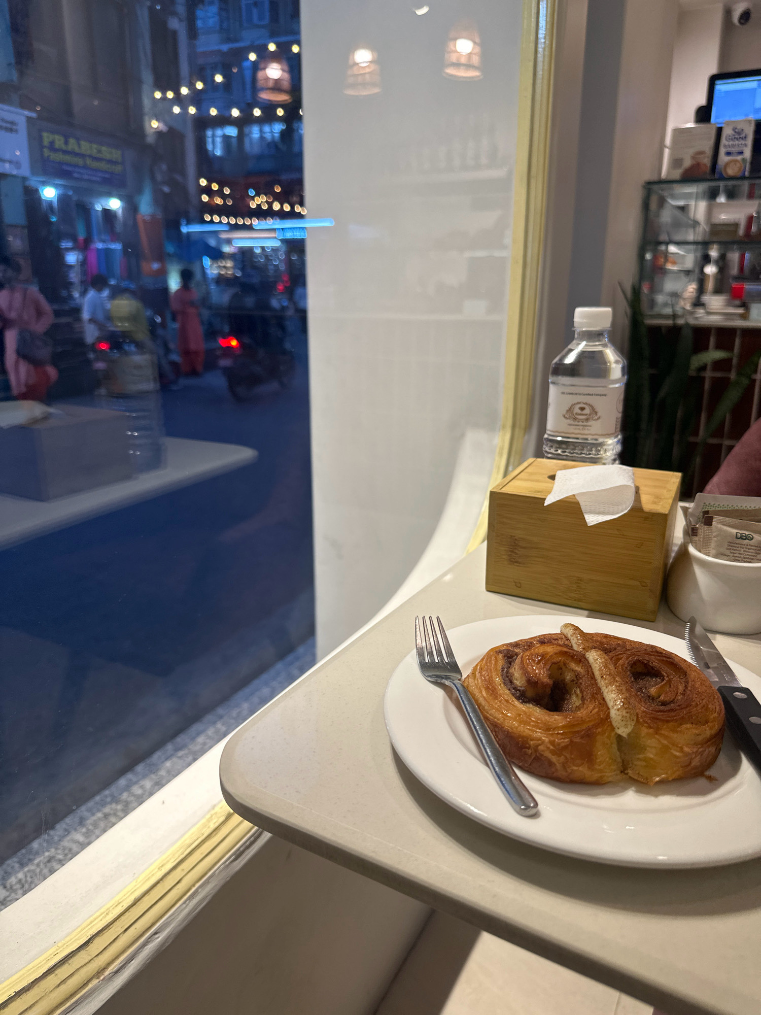 This media file shows a close-up view of a pastry on a white plate, positioned on a table near a window. The pastry appears to be a cinnamon roll or a similar baked good, with visible swirls of cinnamon and icing. A fork rests to the left of the plate, and a knife is placed to the right.
In the background, through the window, a street scene is visible. The scene appears to be a market or shopping area, with several people walking and motorcycles on the road. Strings of lights are hung overhead, suggesting it might be evening or a festive occasion. The reflection in the window shows a blurred view of the interior of a cafe or shop, with shelves and a computer monitor.
A bottle of water and a wooden tissue box are also on the table, near the window. A small white bowl containing sugar packets is visible to the right of the tissue box. One sugar packet has visible text that reads "DBo" and mentions "Manufactured & Packaged by DBO". The overall setting suggests a cafe with an outdoor view. The time of day is likely evening, indicated by the artificial lights in the background. The location context is Kathmandu Metropolitan City, Nepal.
