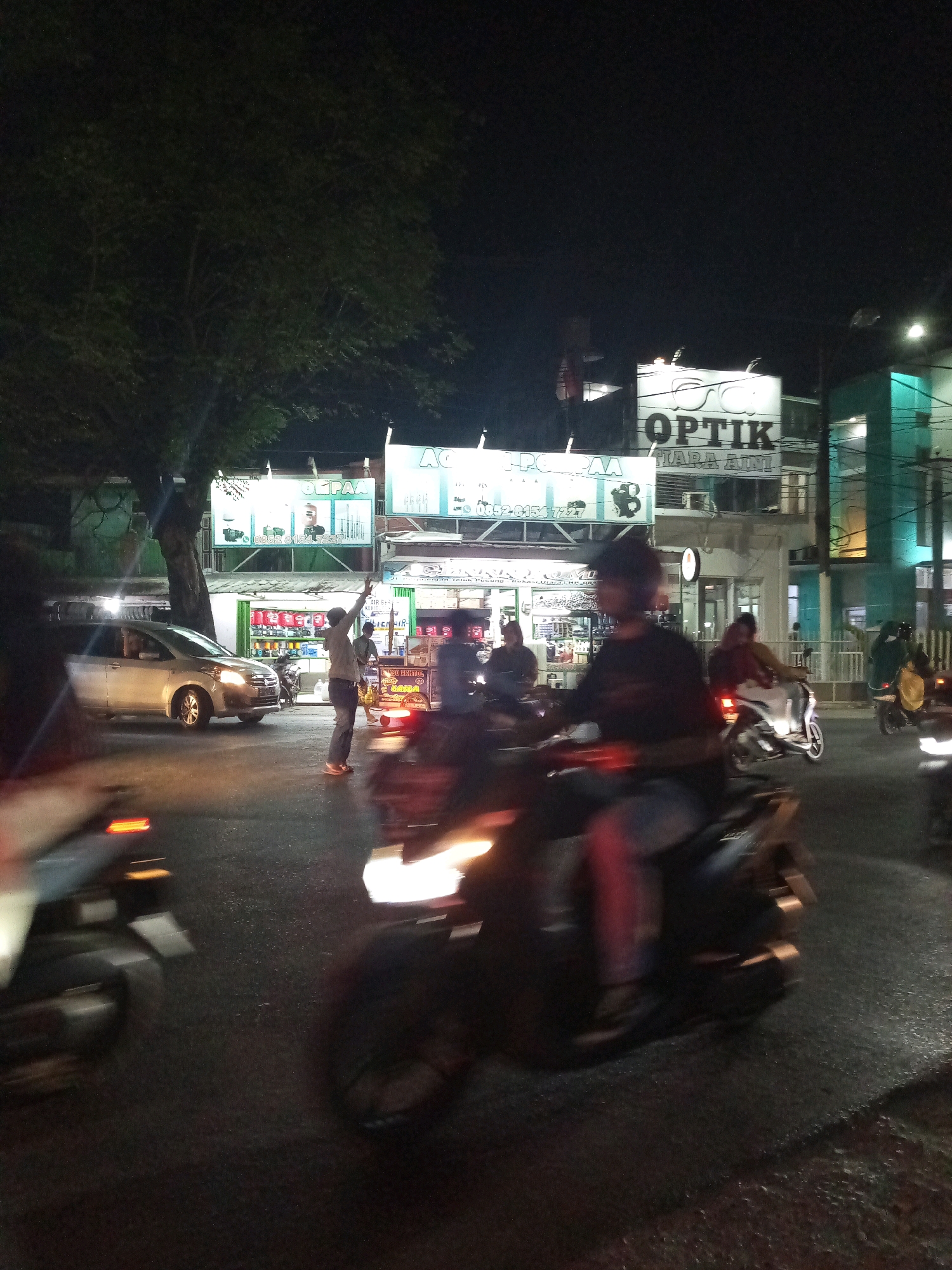 The image depicts a nighttime street scene in Bekasi, Indonesia, likely on a road with commercial establishments lining one side. In the foreground, a dark motorcycle with a rider is moving from left to right, its headlight illuminated and casting light on the wet asphalt. To the left, a silver car is also in motion, its headlights on. In the middle ground, a man is standing on the side of the road, facing away from the camera and raising his right arm, possibly directing traffic or signaling. Behind him, several motorcycles are parked or moving. Various signs are visible on the buildings, including one that reads "OPTİK TIARA ADIM," indicating an optical shop. Other businesses appear to be selling goods, with shelves of products visible through open storefronts. The overall impression is of a busy urban environment at night, with traffic and pedestrian activity.