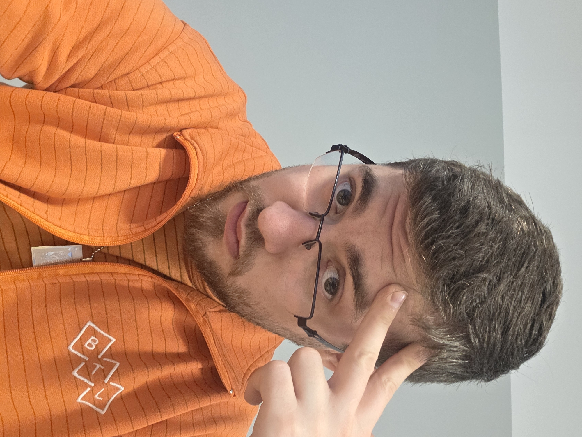 A person wearing glasses and an orange striped jacket is looking down at the camera. The person is touching their forehead with their finger. The background is a light gray wall. The jacket has a white logo with the letters "BTL" arranged vertically within diamond shapes. The location in Plovdiv, Bulgaria cannot be determined from the image.