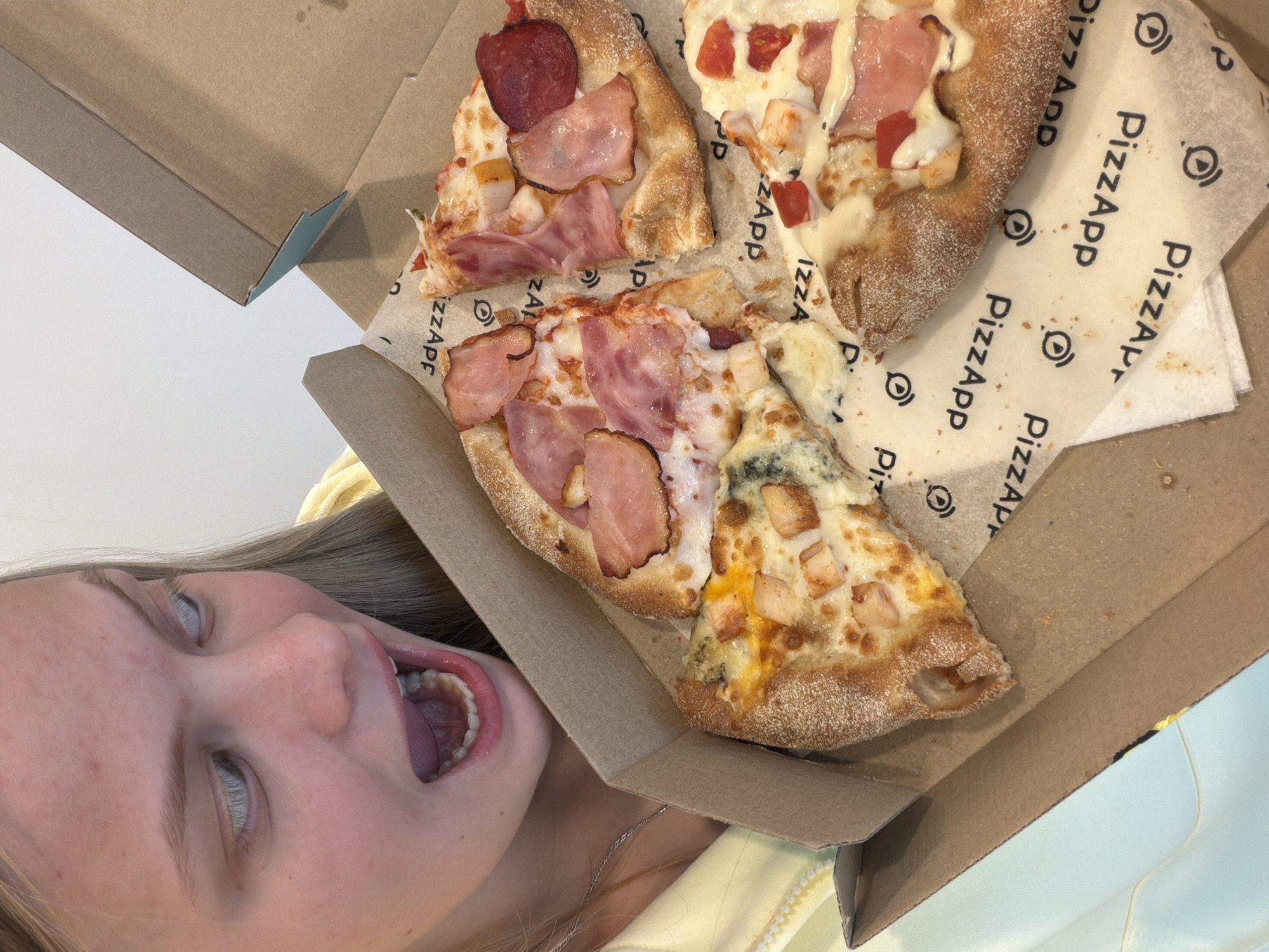 A young fair-skinned person with light hair is positioned at the bottom-left of the image, with their mouth open as if about to consume food. A cardboard pizza box is situated above and to the right of them. Inside the box, three pizza slices are visible. The paper lining the pizza box has a repeating print of the text "PizzApp". The slices have various toppings, including ham and what appears to be pepperoni. The location is not identifiable from the image.