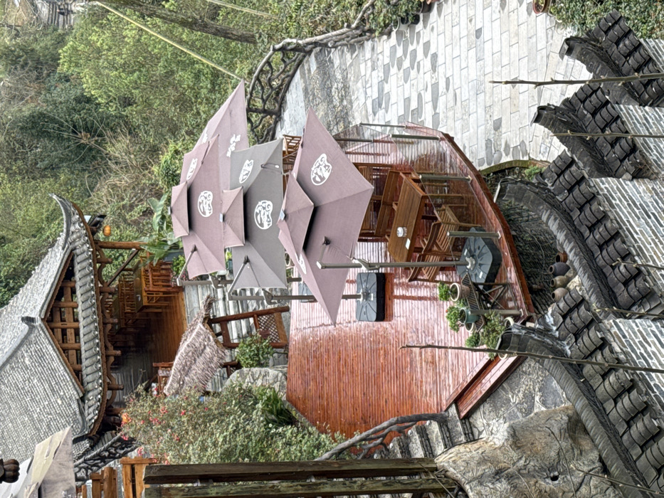 This image depicts a multi-level outdoor dining or recreational area situated on a hillside in Furong, China, under overcast conditions. The scene shows no individuals, actions, or interactions.

In the upper-left, a traditional Chinese-style building with a dark tiled, curved roof and ornate wooden eaves is visible. Its interior features multiple light brown wooden tables and chairs. A single red lantern hangs from the eaves.

Below and to the right of this building, a large, reddish-brown wooden deck extends outwards, appearing wet, suggesting recent rain. This deck is furnished with four unoccupied outdoor seating arrangements. Each consists of a brown, dual-tiered umbrella with a white circular logo, supported by a dark base, shading a rectangular wooden table and several matching wooden chairs. One table contains a white ashtray. Near the edge of the deck, a metal stand holds several potted plants, including bonsai-like specimens. A metal railing encircles the deck's perimeter.

Stone steps with a wooden handrail ascend from the left side of the deck towards the upper building. Further left, a smaller, thatched-roof awning covers another set of stone steps.

To the right of the wooden deck, a winding pathway made of light grey and white patterned stone tiles is visible. This path is bordered by a rustic, branch-like wooden balustrade. Dense green foliage and trees cover the background and parts of the left side of the scene.

In the foreground, below the deck, a traditional brick wall with dark tiled eaves is present. Several brown clay pots are arranged on these eaves.