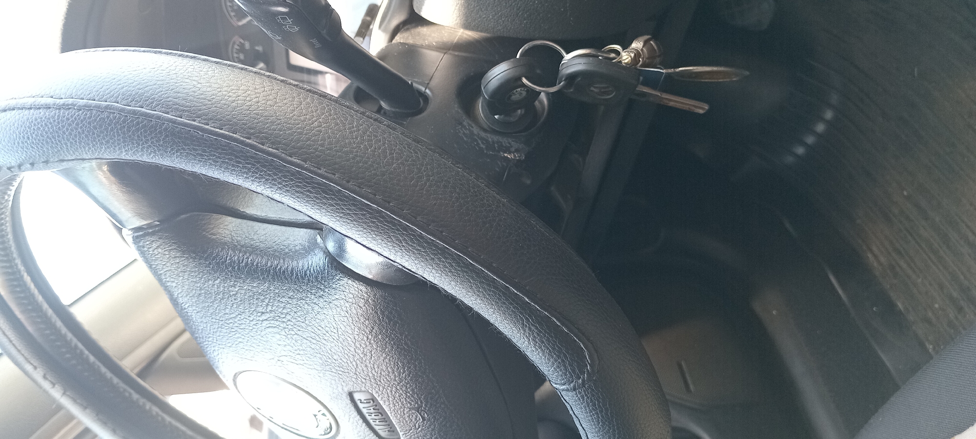 The image displays a close-up view of a car's interior, specifically focusing on the steering wheel and ignition. The steering wheel is black and appears to be covered with a textured material. On the left side of the steering wheel, the word "AIRBAG" is visible. To the right of the steering wheel, an ignition key with a keychain is inserted into the ignition slot. The keychain has a metallic ring and a black fob, which appears to have a car logo on it, possibly Volkswagen based on the visible symbol on one of the keys. A turn signal lever is also partially visible to the right of the steering wheel. The background shows parts of the car's dashboard and the floor mats, which are dark and ribbed. There are no people visible in the image. The lighting suggests it is daytime, with light coming from the right, casting some shadows. The location of Koviahy, Ukraine is not identifiable from the visual information provided in the image.