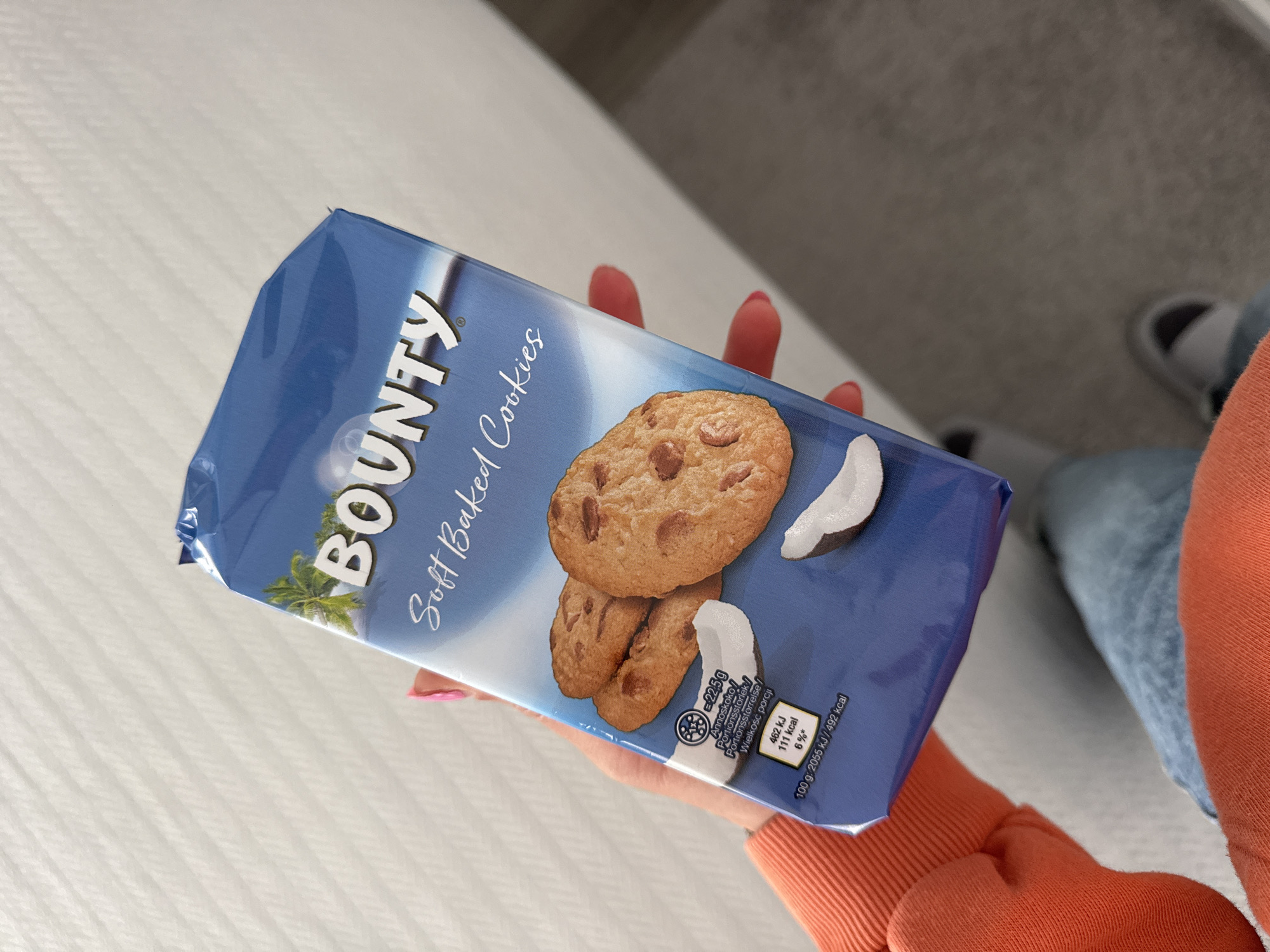 A hand holds a package of Bounty Soft Baked Cookies. The packaging is blue and features images of cookies and coconut. Nutritional information is visible on the package, including details on calories and portion size. The background includes a bed and part of a person wearing an orange sweater. It is impossible to tell if the location is in the city of Blagoevgrad, Bulgaria.