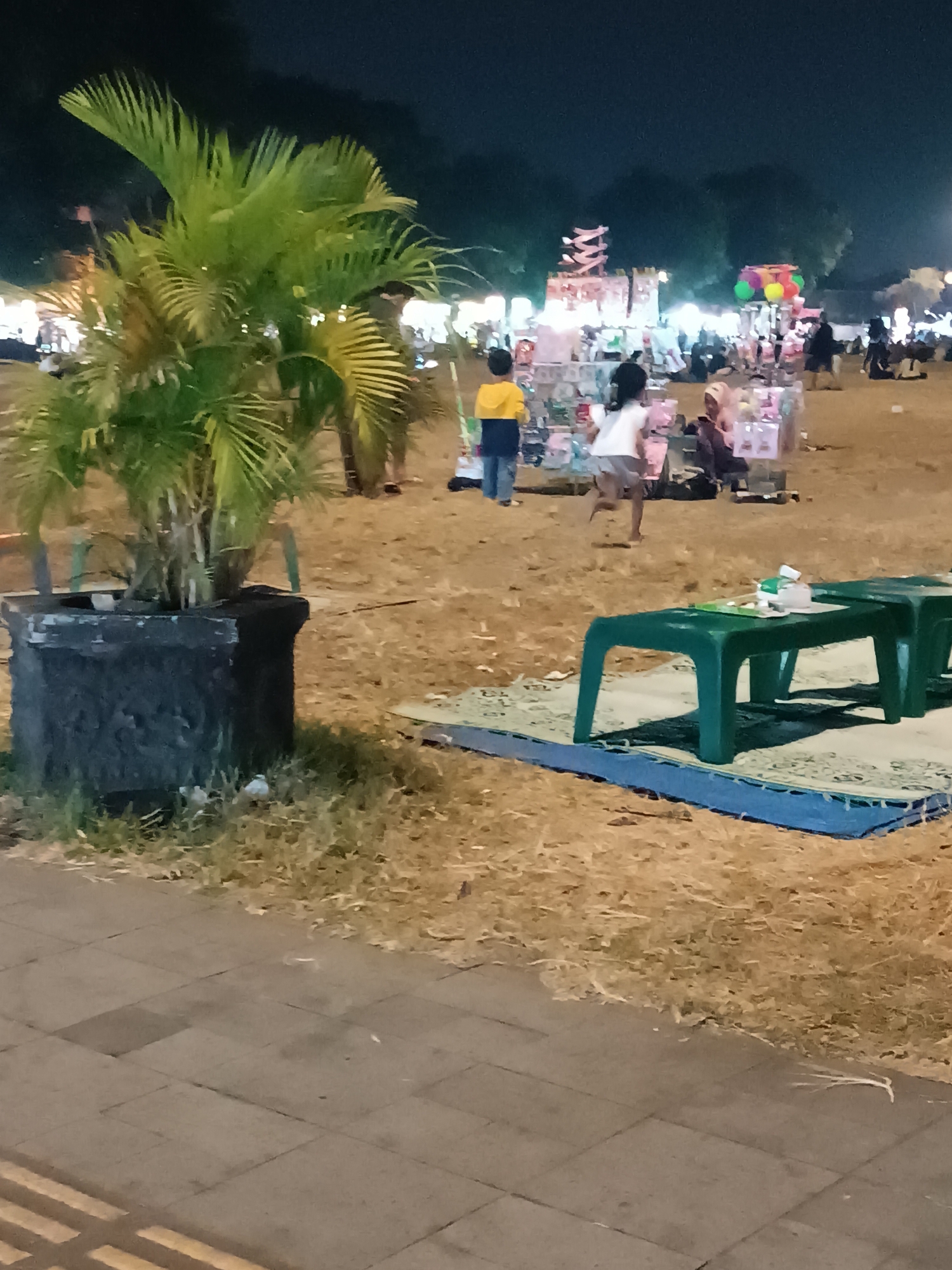 This is a nighttime outdoor scene in a city park in Yogyakarta, Indonesia. The foreground features a large potted palm tree in a dark, weathered planter. To the right, a blue tarp is laid on dry grass, topped with a woven mat, on which are two green plastic benches with some white items on them. In the background, several stalls are set up with lights, displaying goods. A variety of people are milling around the stalls, some standing and some sitting. Two children are visible, one in a yellow top and blue pants, and another in a white top and shorts. The ground is a mix of dry grass and some paved areas. The overall atmosphere suggests a night market or a similar community gathering.