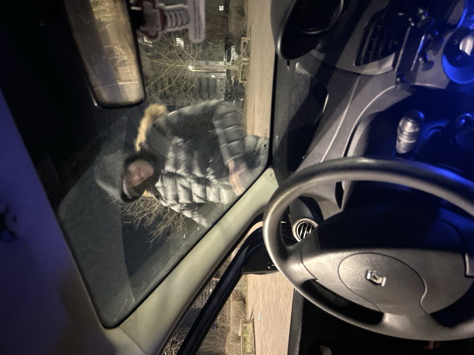 The image depicts a nighttime scene viewed from inside a vehicle in Ostwald, France. The vehicle's interior shows a black, three-spoke steering wheel with a Renault logo on the left, and a dark dashboard with some blue illuminated controls and air vents. A rearview mirror hangs from the windshield, displaying reflections and an attached air freshener.
Outside the passenger side of the vehicle, a single individual is visible. This person is wearing a dark, puffy winter jacket with a fur-lined hood pulled over their head, beneath which a dark cap is visible. Their face is partially obscured by the hood and cap, but their mouth is open in what appears to be a smile or smirk, revealing teeth. Their left arm is slightly raised. The individual is looking directly into the vehicle.
The exterior environment is dimly lit by ambient light sources. In the background, bare deciduous trees are visible, along with a paved surface, possibly a parking lot or street. To the left, a light-colored wooden bench is present. Further in the distance, parked cars and parts of multi-story buildings can be discerned. The sky is dark, indicating night.