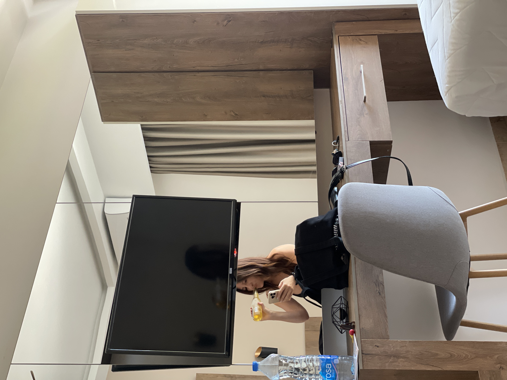 A woman is reflected in a mirror in a room, holding a bottle and a phone. The room appears to be a hotel room in Donji Štoj, Montenegro. A large television is mounted on the wall above a mirrored surface. A wooden dresser or vanity with a drawer is positioned beneath the mirror. A grey upholstered chair with wooden legs sits in front of the dresser. To the left of the dresser, a bottle of water and a black backpack are visible. To the right, a portion of a white bed is in view. The walls are a neutral color, and beige curtains are drawn on the right side of the mirror. An air conditioning unit is mounted on the wall above the television.