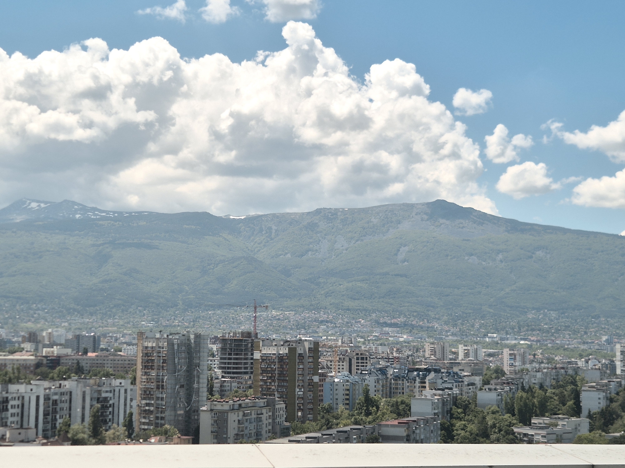 A medium-distance, eye-level shot shows a cityscape, with a mountain range in the background and a predominantly white or light-colored ledge or wall at the bottom of the frame.
In the sky, puffy white clouds contrast with a bright blue expanse. The mountain range is green, suggesting lush vegetation, with some snow-capped peaks visible.
The cityscape features a mix of low-rise and high-rise buildings, with a few construction cranes indicating ongoing development. The architecture seems a mix of Soviet-era apartment blocks and more modern buildings. Trees are interspersed among the buildings.
Given the description, the location is Sofia, Bulgaria, with the mountain range being Vitosha Mountain.