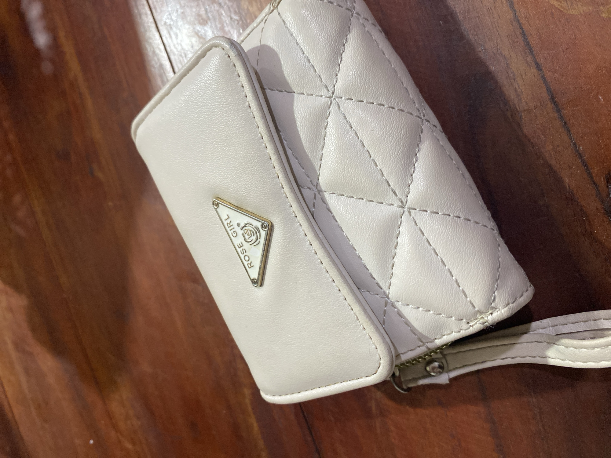 A cream-colored wallet with a quilted diamond pattern is placed on a dark wood surface. The wallet has a flap closure with a triangular gold-toned emblem that reads "ROSE GIRL" with a rose graphic. A wrist strap is attached to the side of the wallet. The wood surface appears to be a table or desk with visible grain and shadows. The lighting suggests an indoor environment.