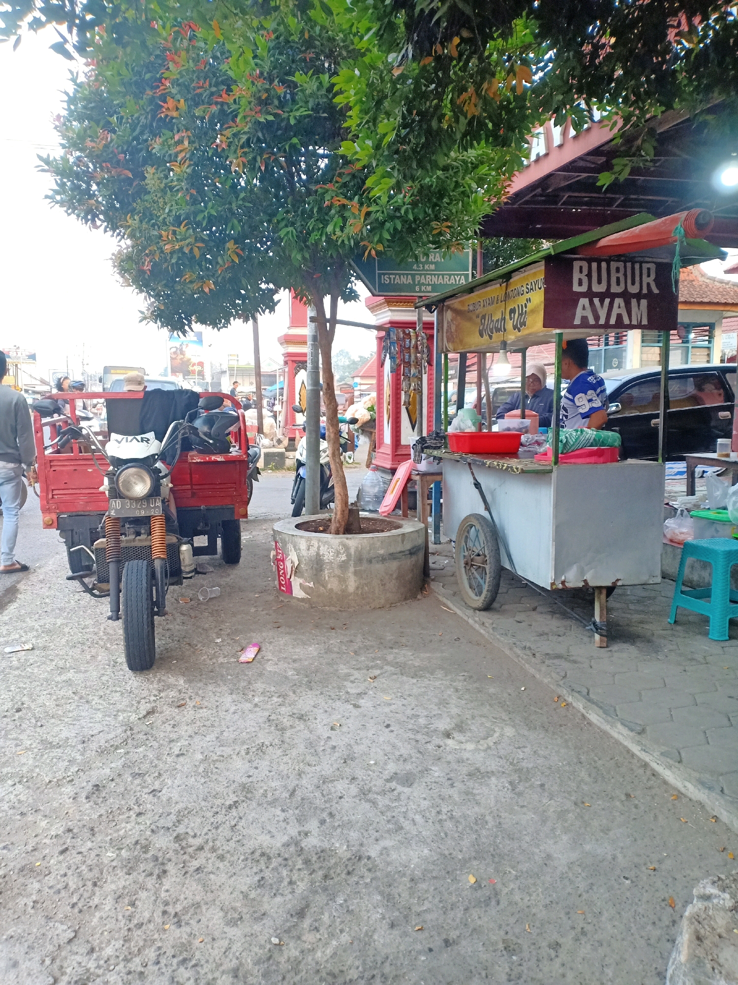 In Mojokerto, Indonesia, a street scene features a food cart with a "BUBUR AYAM" (chicken porridge) sign. The cart is mobile, mounted on wheels, and displays various food items and containers. Two individuals are present near the cart; one is serving food, while the other appears to be a customer. A red tricycle, laden with items, is parked to the left of the food cart. The background shows a building with traditional Indonesian architectural elements, including red columns and a dark-tiled roof. Trees and foliage are visible, with one prominent tree casting shade over the scene. The ground is paved with concrete, and scattered debris indicates a public area. The lighting suggests it is daytime, possibly late afternoon, given the soft shadows.