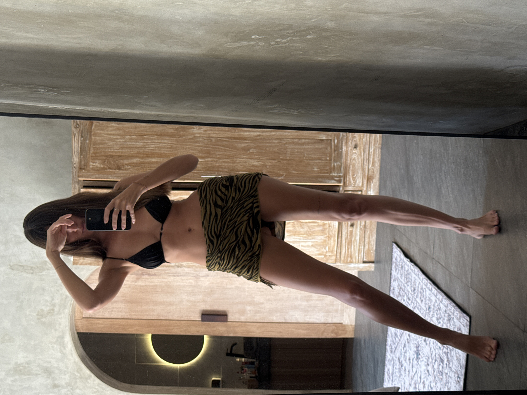 A young woman stands, reflected in a large mirror, taking a full-body selfie with a smartphone. She is wearing a black bikini top and a green and black tiger-print skirt. Her long, dark hair is down, and she is holding the phone with her right hand while her left hand is raised near her face.
The setting appears to be an indoor space, likely a modern bathroom or dressing area, with elements consistent with a tropical or contemporary aesthetic found in locations like North Kuta, Indonesia. The background, as seen in the reflection, features a warm-toned wooden cabinet or door, alongside walls and a floor with a concrete or stone texture.
In the lower left of the reflection, a striking design element is visible: a semi-circular alcove with a soft, warm glowing light, possibly part of a vanity area with dark, sleek countertops and various bottles. On the floor in the foreground, a patterned rug is visible. The lighting is artificial and ambient, creating a clean and modern atmosphere. No legible text is visible in the image.