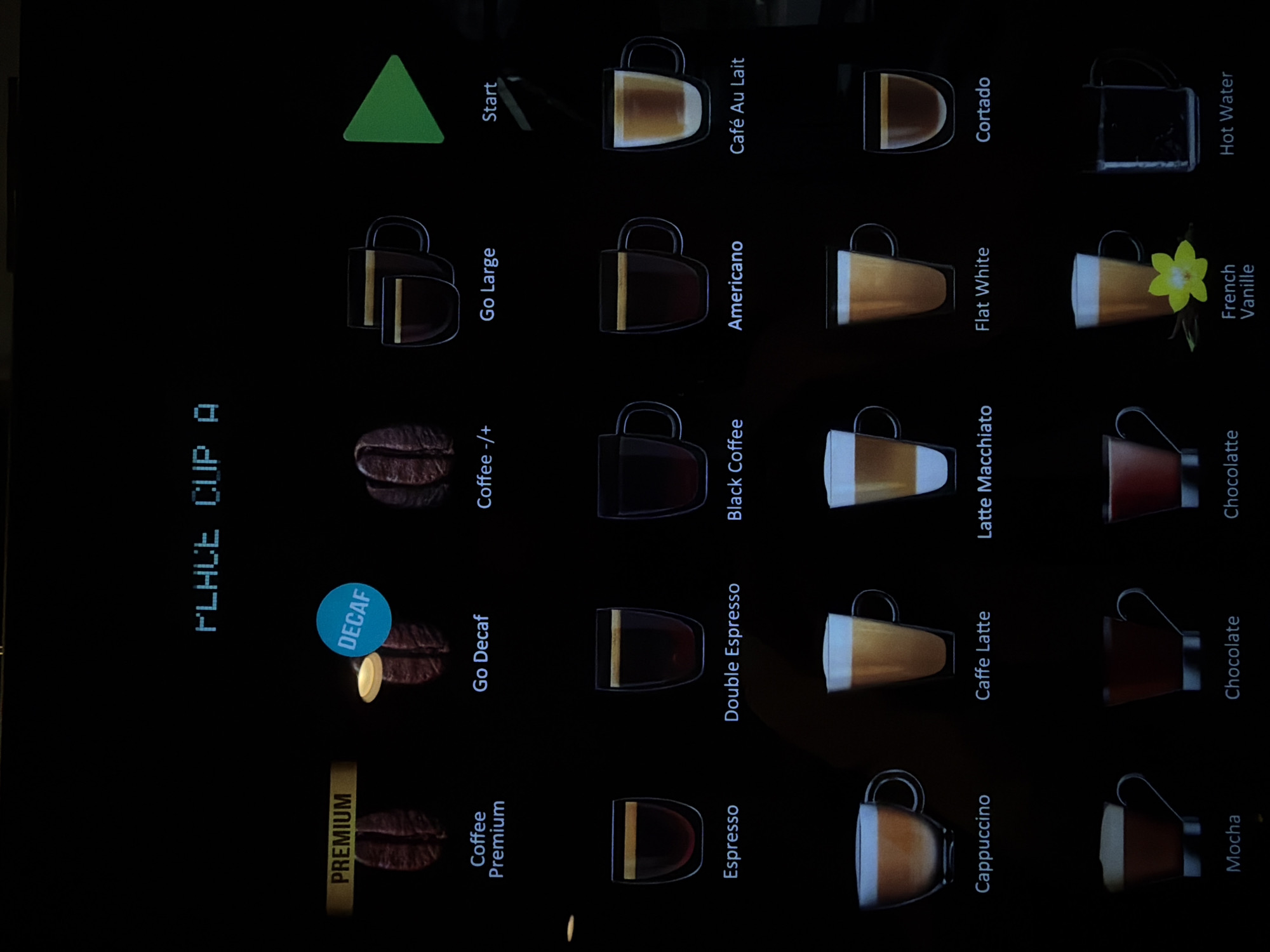 This image shows a digital display of a coffee machine menu. The display is black with white and blue text and images. At the top, the text "PLACE CUP A" is visible. Below this, there are several coffee options presented with corresponding images. From left to right and top to bottom, the options are:
Top row:
- "PREMIUM" text above a coffee bean image, labeled "Coffee Premium"
- A light shining on a coffee bean image, labeled "DECAF" and "Go Decaf"
- A coffee bean image, labeled "Coffee -/+""
- Two stacked coffee cups, labeled "Go Large"
- A green play button, labeled "Start"
Second row:
- A glass cup with dark liquid, labeled "Espresso"
- A glass cup with dark liquid, labeled "Double Espresso"
- A glass cup with dark liquid, labeled "Black Coffee"
- A glass cup with dark liquid, labeled "Americano"
- A glass cup with light brown liquid, labeled "Café Au Lait"
Third row:
- A glass cup with light brown liquid and foam, labeled "Cappuccino"
- A tall glass cup with light brown liquid and foam, labeled "Caffe Latte"
- A tall glass cup with layered light brown liquid and foam, labeled "Latte Macchiato"
- A glass cup with light brown liquid, labeled "Flat White"
- A glass cup with dark liquid and foam, labeled "Cortado"
Fourth row:
- A cup with dark liquid and foam, labeled "Mocha"
- A cup with dark liquid, labeled "Chocolate"
- A cup with dark liquid and foam, labeled "Chocolatte"
- A glass with a yellow flower and a cup with light brown liquid, labeled "French Vanille"
- A glass cup filled with clear liquid, labeled "Hot Water"
The image appears to be taken indoors, likely in a commercial setting where a coffee machine is present. There is no information in the image that allows for determination of its location in the Town of East Hampton, United States of America. The scene is a digital interface on a screen, with no people or specific environmental details visible beyond the display itself.
