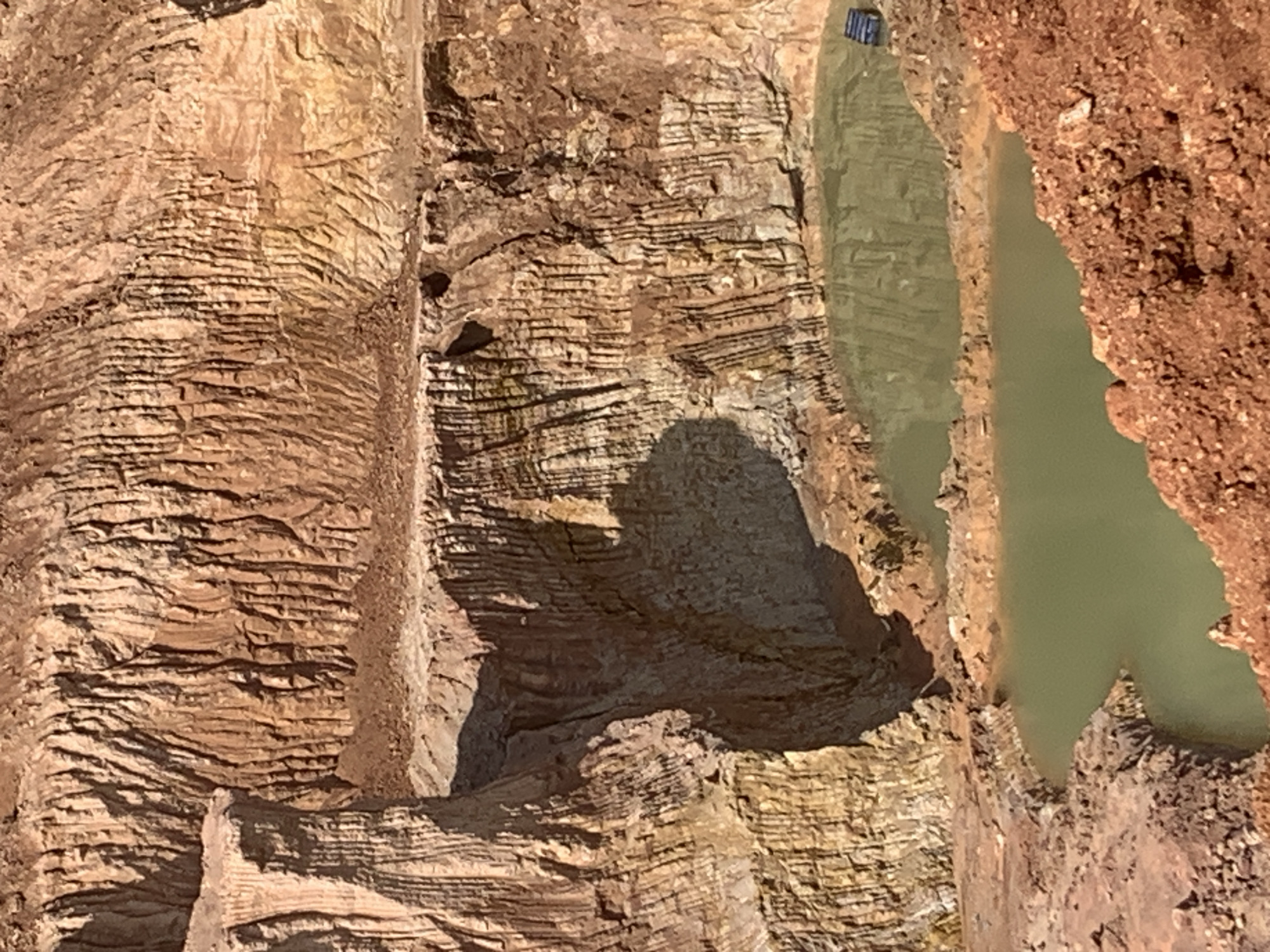 The image shows a quarry in Kwang, Nigeria. The quarry is a large pit in the ground with steep, layered walls of reddish-brown earth and rock. In the bottom of the quarry are two pools of murky, green water. The water reflects the quarry walls and the sky. There is a small, dark blue object, possibly a piece of equipment, floating in the upper pool of water on the right side. The overall scene depicts an active or recently active excavation site.