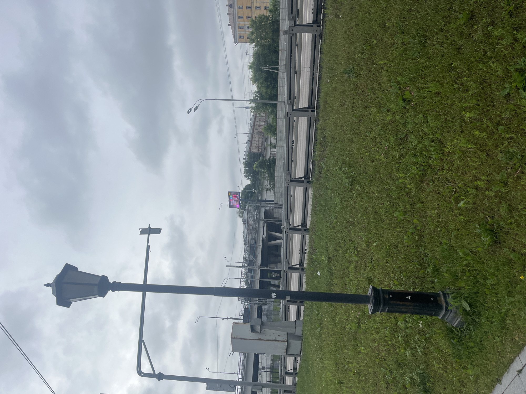 The image depicts a street scene in Moscow, Russia, under an overcast sky. In the foreground, a black lamppost with an ornate base stands on a patch of overgrown green grass. To the left of the lamppost, a large grey utility box is visible. Behind the grass and the utility box, a multi-level highway overpass dominates the midground. The overpass structure is a mix of concrete and metal, with visible scaffolding on some sections, suggesting ongoing construction or maintenance. A grey metal barrier separates the highway from the foreground. In the distance, several buildings, including a yellow residential building, line the horizon. Power lines are strung across the sky. A billboard with a pink advertisement is mounted on a structure near the overpass on the left.