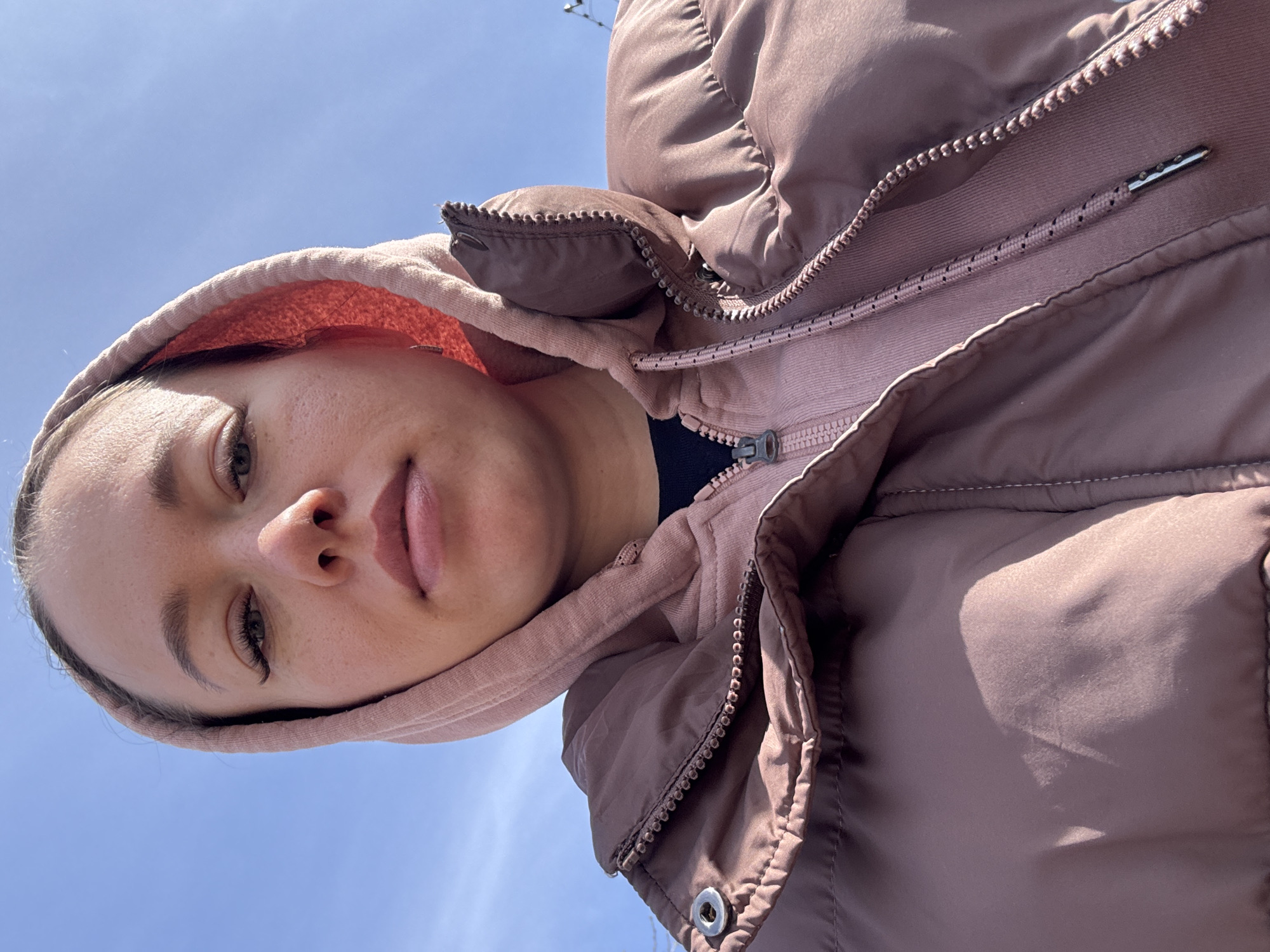A selfie perspective shows a person, from approximately the chest up, wearing a pink hooded sweatshirt under a brown, puffy jacket. The sky is visible in the background. There are no indications of the city of Ferma, Russia.