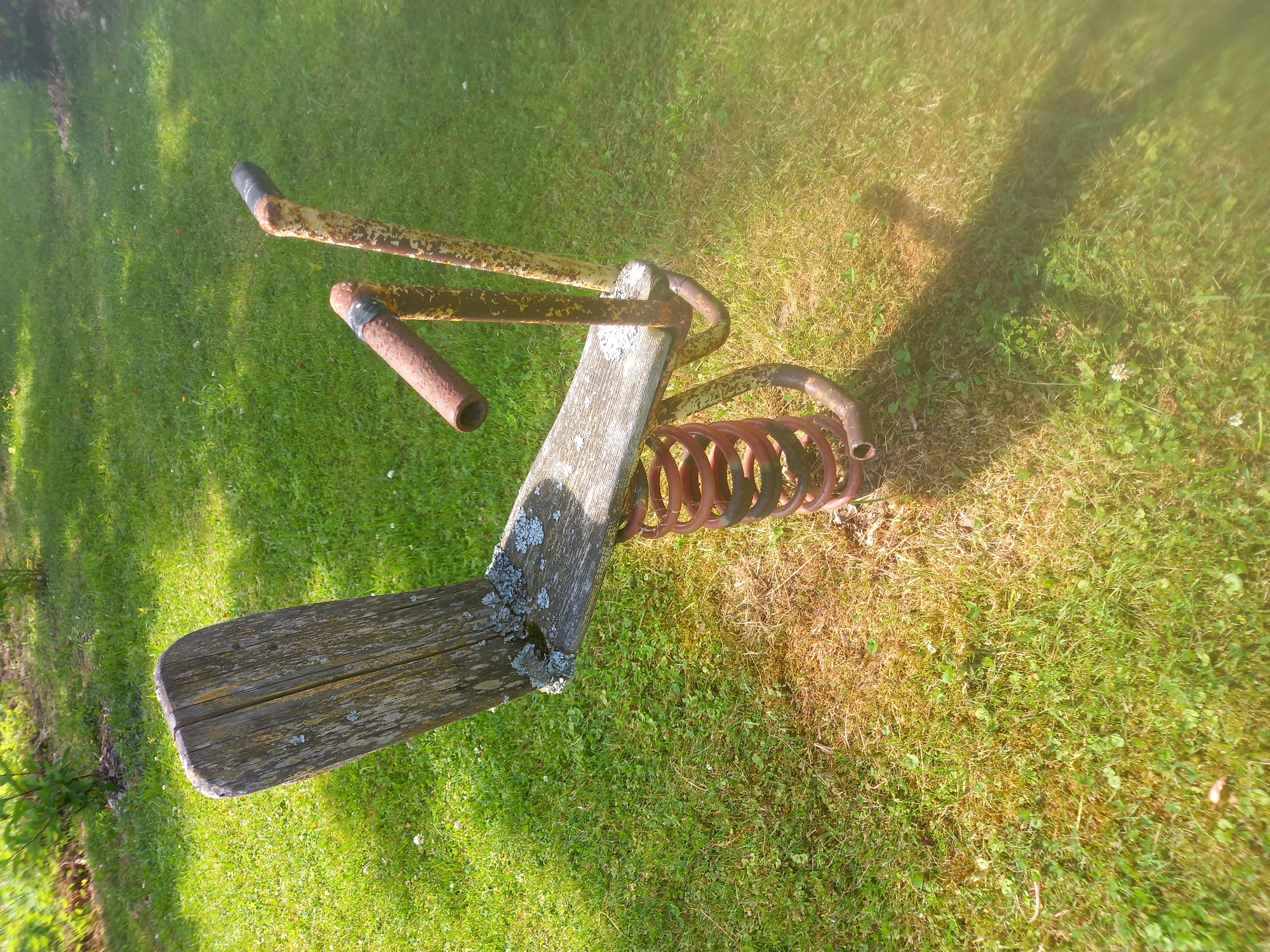 An aged, wooden-seated spring rider, weathered and showing signs of lichen growth, is situated on a patchy lawn with areas of dry grass. The metal structure supporting the seat and handlebars is rusty, painted yellow and red, with some black tape on the handlebars. The rider is positioned in a grassy area, with shadows from nearby foliage cast across the lawn. The overall scene suggests an outdoor recreational space, possibly a park or a backyard, located in Adutiškis, Lithuania.
