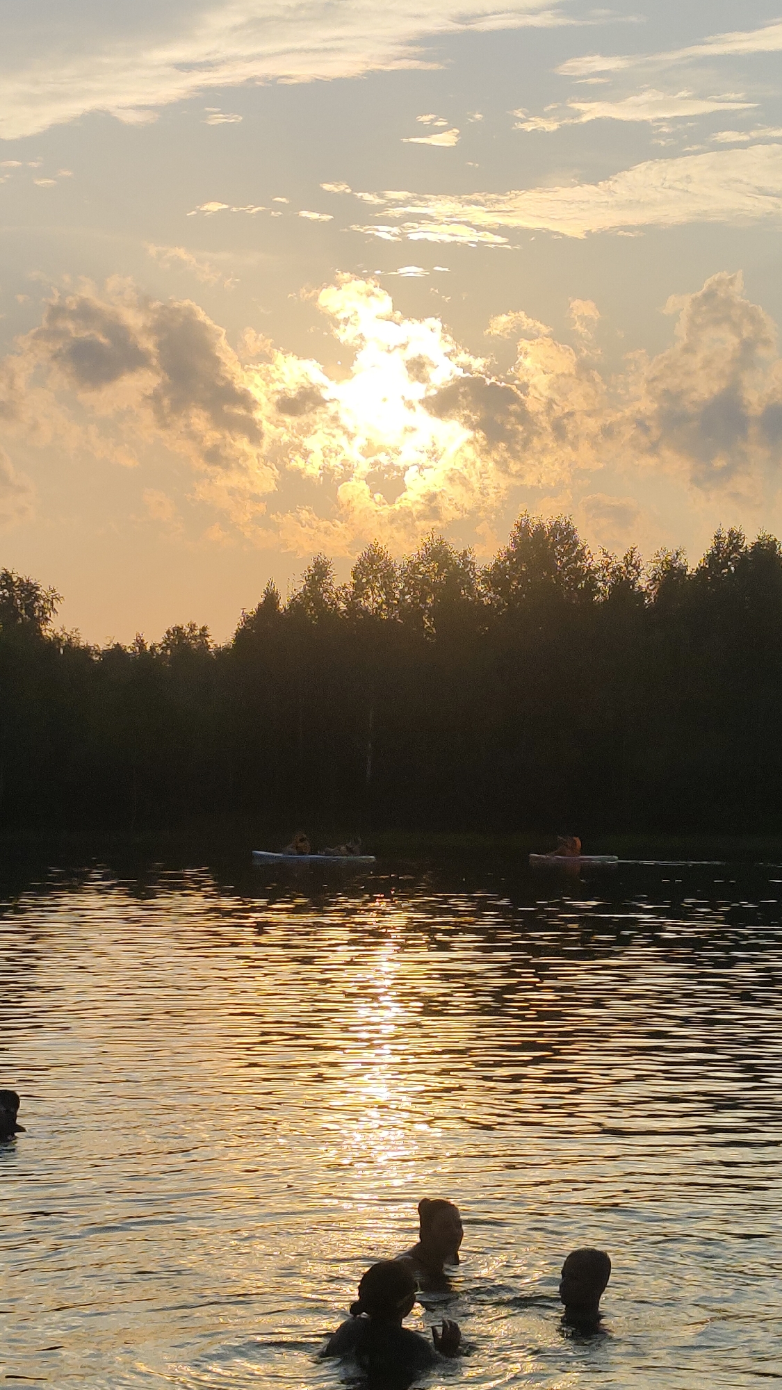 A vertical shot captures a sunset over a body of water in Terent'yevo, Russia. The sky is filled with dramatic clouds illuminated by the setting sun, which peeks through the cloud cover. In the distance, a dense line of dark green trees forms the treeline along the water's edge. On the water, two paddleboards are visible, each with a person on it. Closer to the foreground, several people are swimming in the water, with their heads and shoulders visible above the surface. The water reflects the warm, golden light of the sunset, creating rippling patterns across its surface.