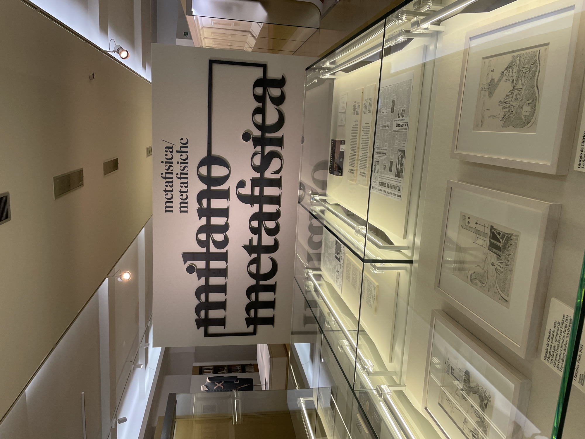 The image shows a display in Milan, Italy, likely in a museum or gallery. A vertical sign reading "milano metafisica / metafisiche" in bold, black letters stands prominently in the foreground. To the right of the sign, framed artworks, including drawings and news clippings, are displayed inside a glass case. The lighting inside the case illuminates the pieces. Artwork details are visible, referencing Giorgio de Chirico and his "Mythologie" series. The background suggests a gallery setting, with architectural details and additional exhibits. The overall impression is that of an exhibition focused on metaphysical art in Milan.