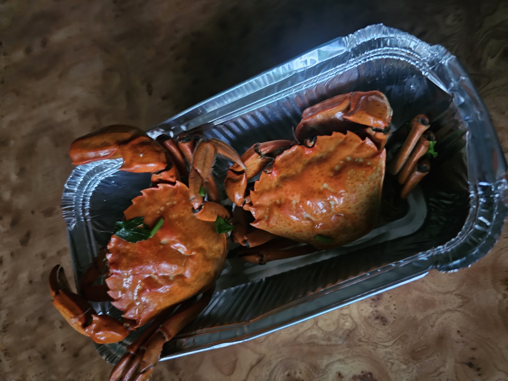 In Skadovsk, Ukraine, two cooked crabs are presented in a disposable aluminum foil tray. The crabs are a vibrant orange-red color, indicative of being steamed or boiled, and are garnished with a few sprigs of green herbs. They are arranged with their legs and claws splayed, filling the majority of the tray. The tray itself is placed on a wooden surface with a distinct grain pattern. The lighting suggests an indoor setting, possibly a kitchen or dining area.
