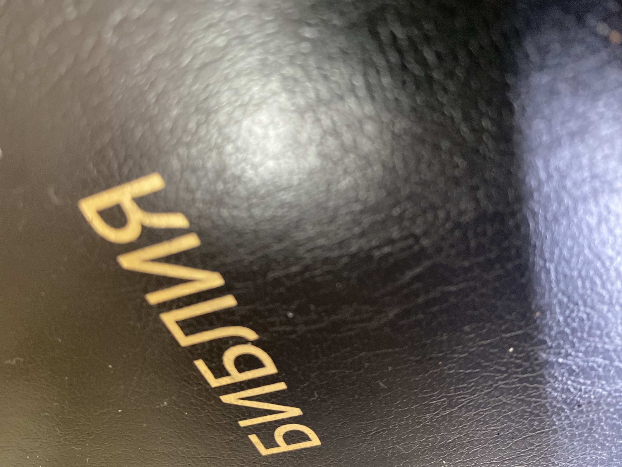 The image shows a close-up of a black, textured surface with the word "БИБЛИЯ" (Bible) printed in gold lettering. The surface appears to be a cover of a book, possibly a bible. The letters are large and slightly blurry due to the close proximity of the camera. The text is rotated counterclockwise. No other details about the scene or location in Ol'khovskoye, Russia can be determined from the image.