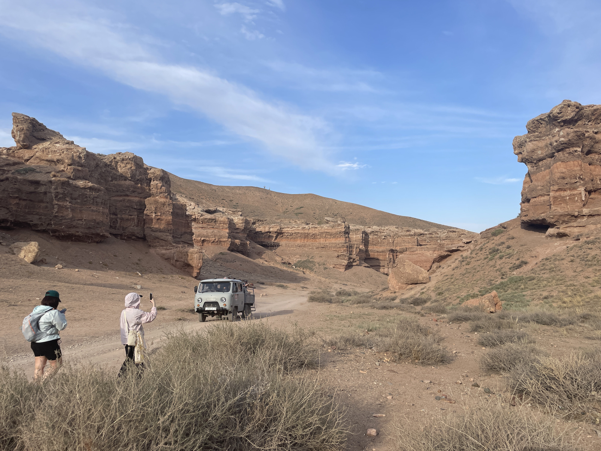 In Tamerlik, Kazakhstan, two individuals are captured mid-stride on a dirt road within a canyon. The person on the left, wearing a green cap, blue jacket, and black shorts, appears to be looking at a phone. To their right, another person in a light pink hooded jacket and dark pants holds up a phone, possibly taking a picture. Ahead of them on the road, a light gray UAZ van, a utilitarian vehicle often used in off-road conditions, is parked or moving slowly. The canyon walls, composed of layered reddish-brown rock, rise on either side, with sparse, dry vegetation dotting the foreground and slopes. The sky above is a clear blue, with wispy white clouds streaking across.