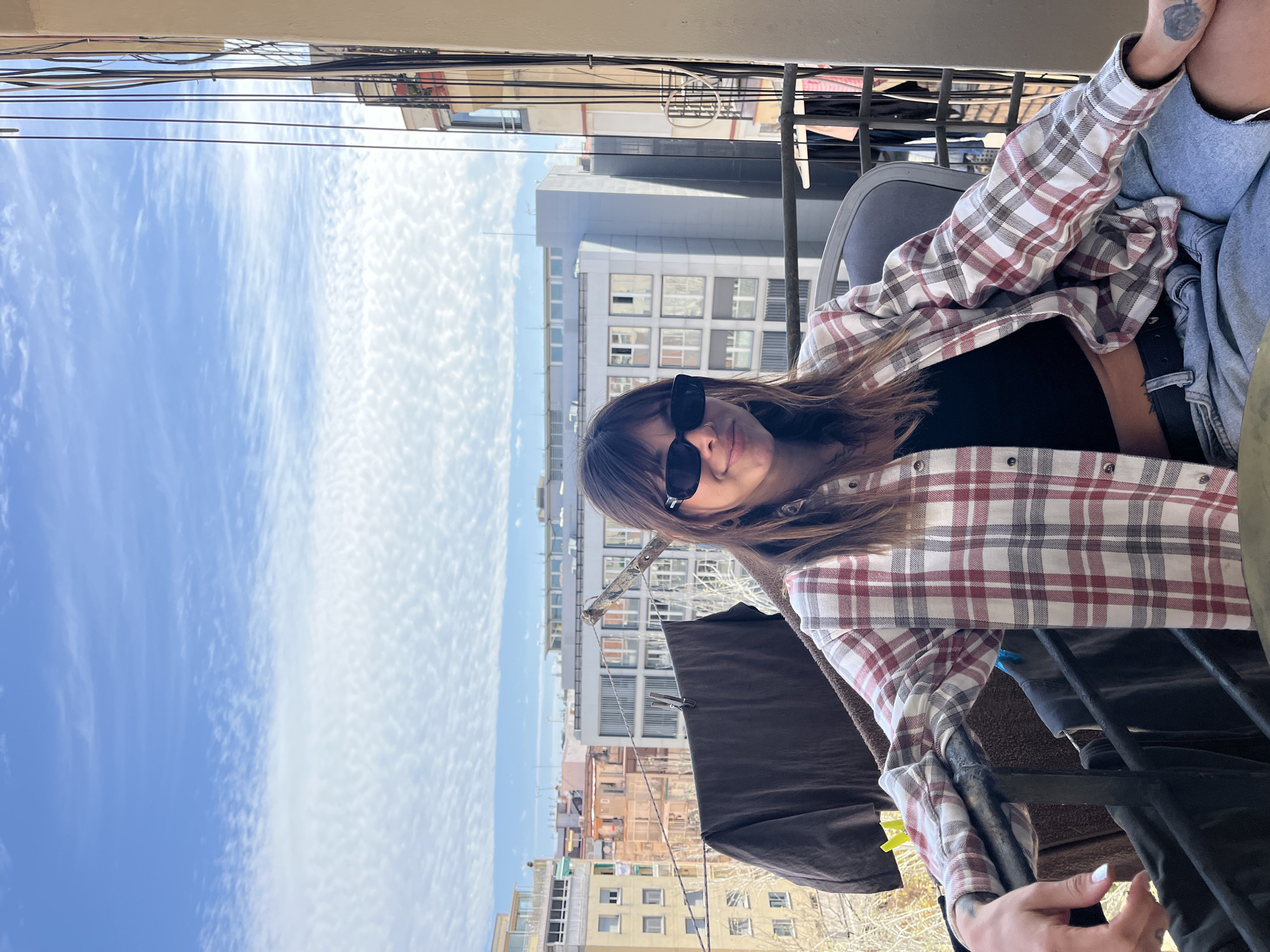 The image captures a young woman relaxing on a balcony, looking directly at the camera with a subtle, relaxed smile. She has brown hair and wears black rectangular sunglasses, a black crop top, and an open plaid flannel shirt featuring a red, white, and dark gray pattern. A small, dark tattoo is visible on her left forearm, and her fingernails are painted white.
The setting is an urban environment, likely Barcelona, as indicated by the context. In the background, a bright blue sky with scattered, wispy white clouds suggests clear and sunny daytime weather. Below the horizon, a dense cityscape is visible, with buildings of various architectural styles and heights. On the balcony itself, part of a metal railing and a clothesline with a dark fabric (possibly a blanket or towel) and clothespins are discernible, common features of Mediterranean balconies. No visible text is present in the image.
