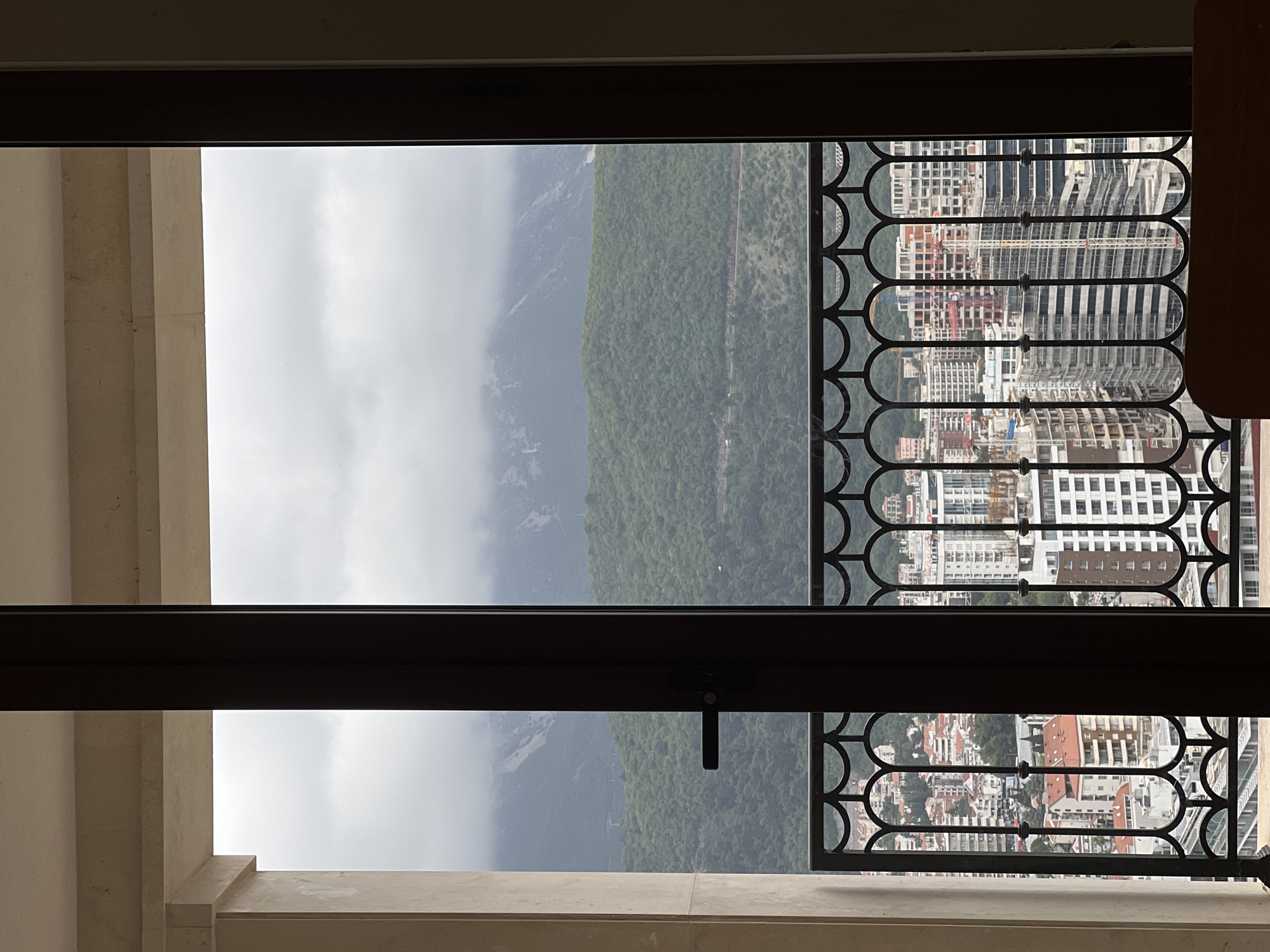 An interior shot through a window or balcony door, offering a view of the landscape outside. The scene includes mountains covered with greenery, visible to the left, and densely packed buildings characteristic of a city environment on the right, with a decorative wrought iron railing in front. The architecture appears modern, with a mix of residential and commercial buildings. The sky is overcast, suggesting a cloudy day. The window or door frame is dark, contrasting with the brighter outdoor scene. The location appears to be Budva, Montenegro, based on the distinctive topography and architecture.