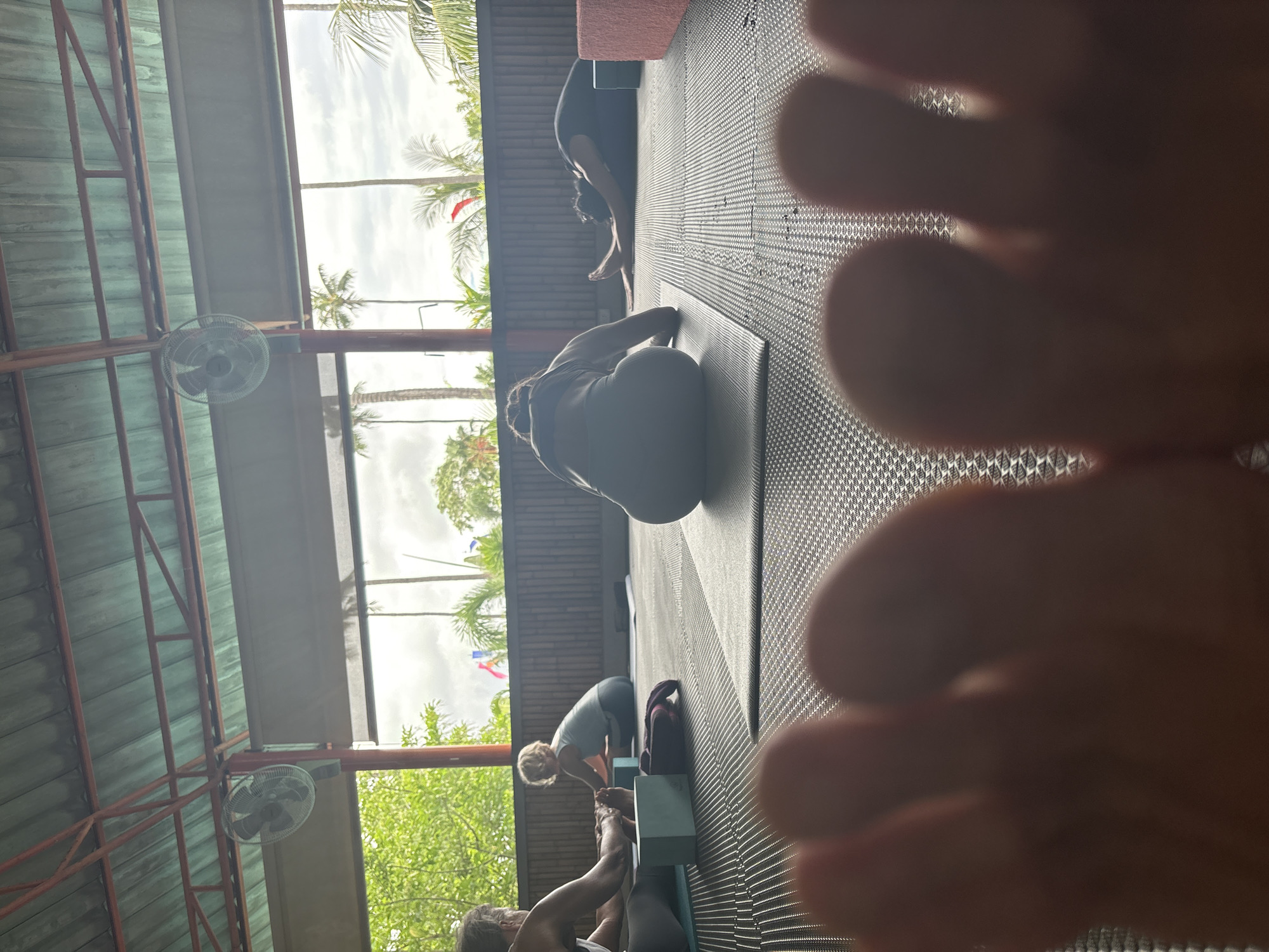 Here is a description of the image:
The image appears to be taken from a very low angle, possibly on the floor of a yoga studio or similar space. The foreground is heavily dominated by out-of-focus feet, seemingly belonging to the person taking the picture. Beyond the feet, a room with multiple people doing yoga is visible. The floor is covered in dark mats, and there are windows along one side of the room, offering a view of palm trees and other greenery outside. The building's roof is visible, with exposed metal beams and ceiling fans. Based on the scenery and the style of the building, it is likely the yoga class is held somewhere in or near Rawai, Thailand.