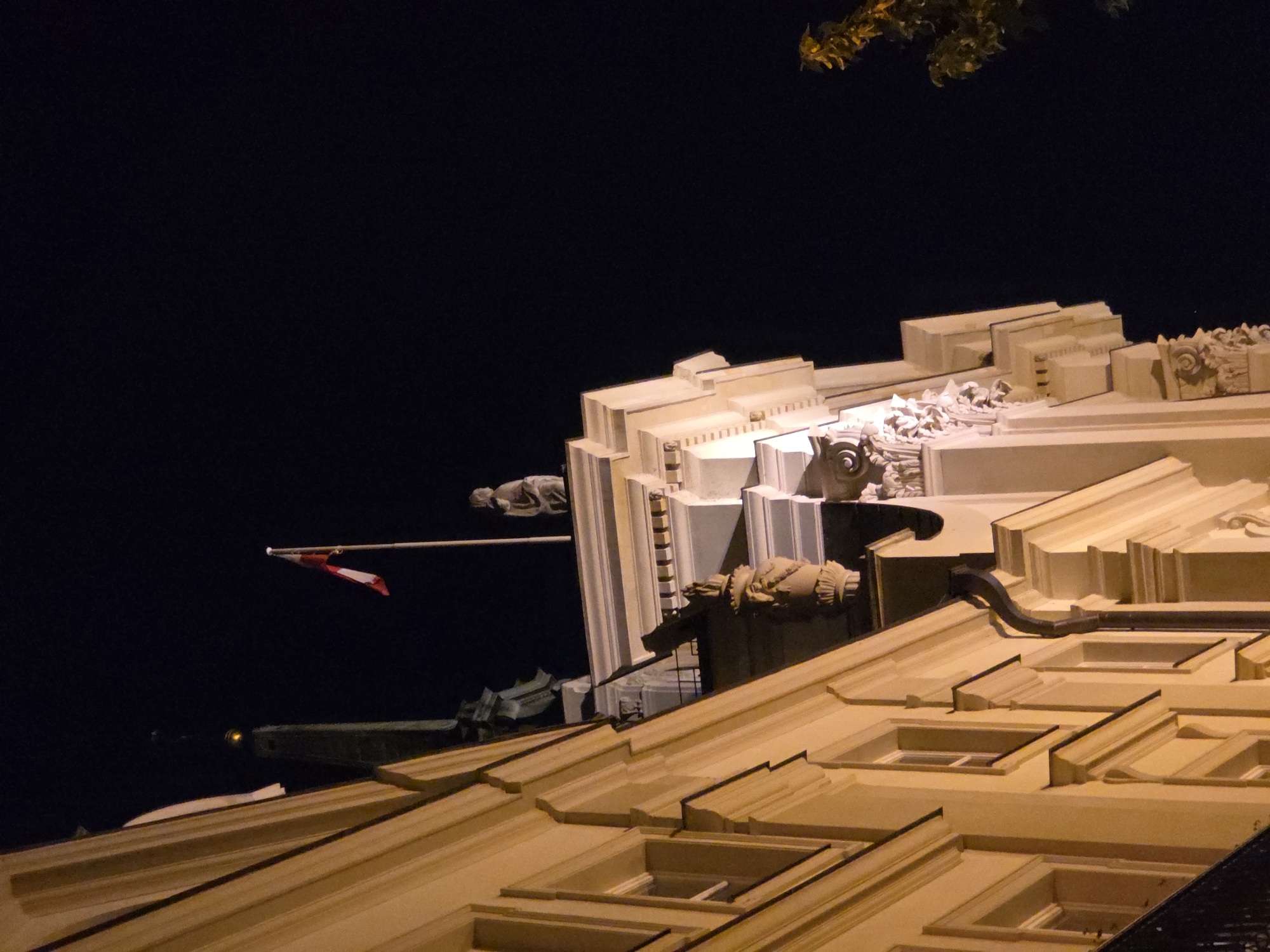Captured at night, the image shows an architectural view of the Polonia Palace Hotel in Warsaw, Poland. The vantage point is low, looking up at the building's ornate facade. The facade is decorated with stone sculptures, gargoyles, and layered geometric ornamentation, typical of early 20th-century hotel design. A Polish flag is visible, projecting from the building, against the dark night sky. The building's roofline is illuminated, highlighting its various levels and architectural details. The scene is dark, with the hotel's architectural elements strongly contrasted against the black sky. In the upper right corner, there is some foliage. The image suggests a carefully composed architectural photograph with a focus on details and the effect of light and shadow.