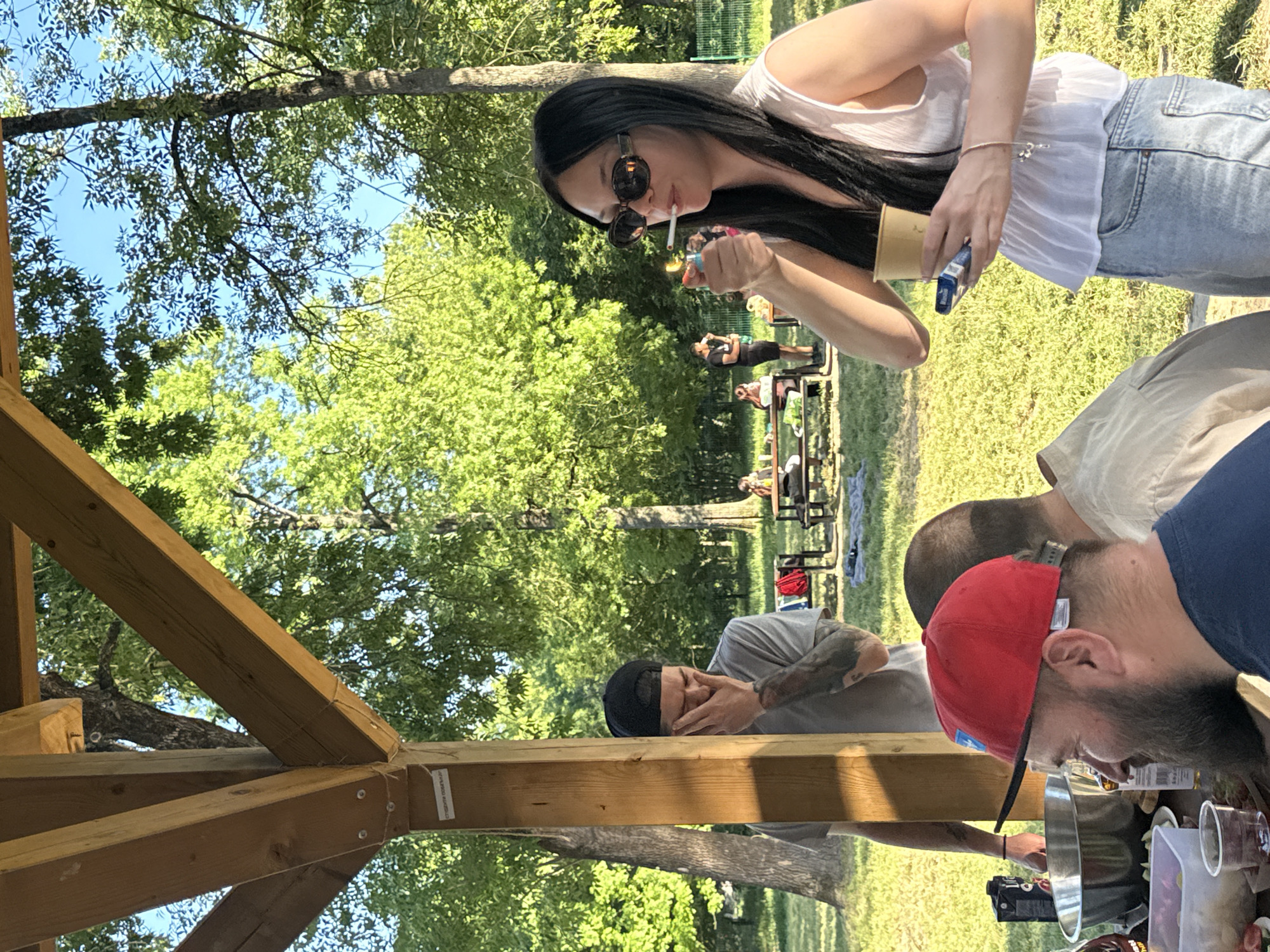 The image shows a group of people in a park setting, possibly in Cukarica Urban Municipality, Serbia, given the prompt's specification. A woman with long dark hair, sunglasses, a white top, and jeans is lighting a cigarette with a lighter. She is holding a cup in her other hand. In the foreground, there are people standing around a table filled with items, including drinks, food, and disposable cups. One man is wearing a red baseball cap and another is wearing a black baseball cap. The scene appears to be outdoors in a grassy area with trees visible in the background. A picnic table with other people is also visible in the background. The photograph is taken from under a wooden structure, possibly a gazebo or a shade structure in the park.