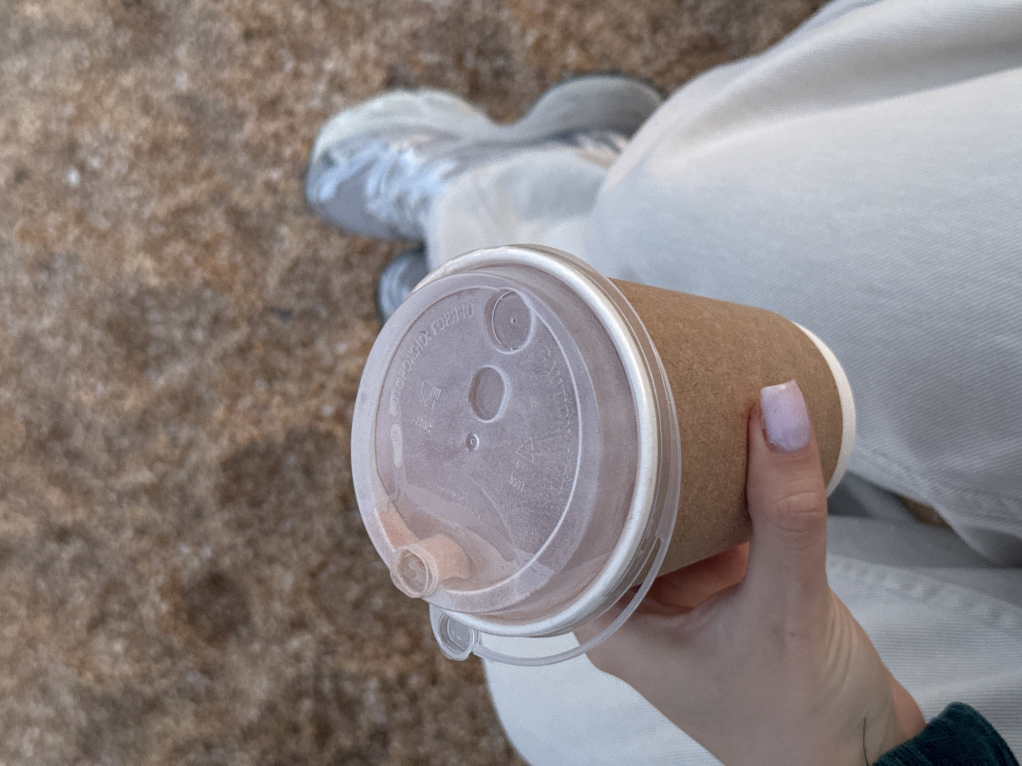A top-down, medium shot depicts a person holding a brown paper coffee cup with a clear plastic lid. The lid has a drinking spout and air holes. The person's hand, with a pale purple fingernail, is visible. The person is wearing light-colored trousers and their shoes are slightly visible in the background. The ground appears to be a textured, brownish surface, possibly sand or concrete. The location is not possible to discern from the image.