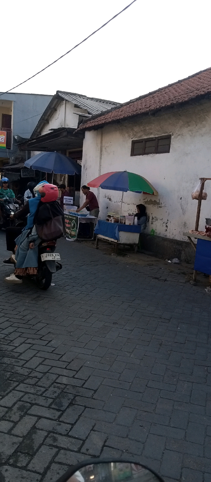 In Wadungasri, Indonesia, a street scene unfolds with a woman on a motorcycle wearing a pink helmet and a blue outer garment, her legs visible beneath a patterned skirt. She is accompanied by a second individual, also seated on the motorcycle. The road is paved with dark, rectangular cobblestones. To the right of the motorcycle, a food stall is set up under a multicolored umbrella, where a man wearing a maroon shirt is attending to a customer. The stall displays various items, including what appears to be food and drinks. Another food stall is visible to the left, partially obscured by a blue umbrella. In the background, buildings with tiled roofs and white walls are present. The sky is a pale, overcast white, suggesting either early morning or late afternoon light.