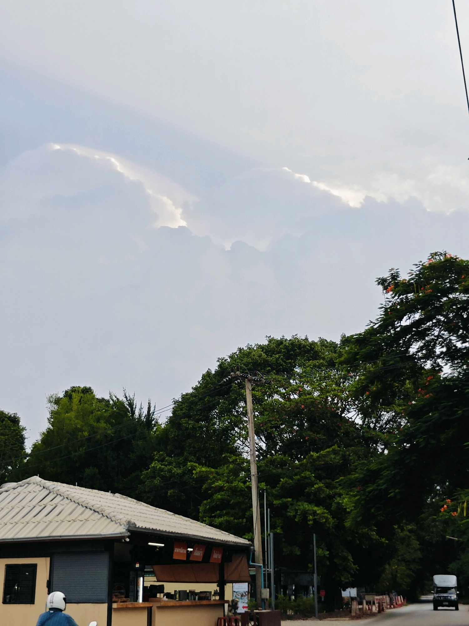 The image depicts a street scene in Mu Si, Thailand, under a cloudy sky. In the foreground, a white tiled roof structure, possibly a small shop or kiosk, is visible on the left. A person wearing a blue shirt and white helmet is riding a motorcycle away from the camera. Behind the structure and to the right, dense green trees line the street, with some trees displaying small orange flowers. A utility pole stands near the left side of the road. Further down the street, a white van is driving away from the camera. Orange and white traffic cones are placed along the side of the road. The sky is overcast with some patches of bright light breaking through the clouds.