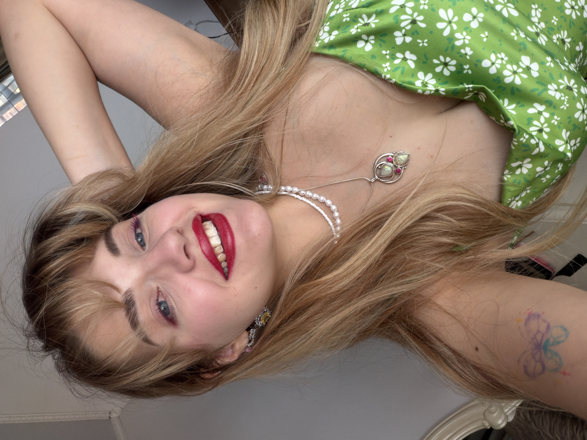 A young woman with blonde hair is lying down and looking at the camera with a smile. She is wearing a green dress with white flowers and a pearl necklace with a pendant. She is also wearing earrings. A butterfly tattoo is visible on her arm. The background appears to be a room with a white wall and a glimpse of a window or mirror. There is no information to indicate the location is in the city of Большая Шильна, Russia.