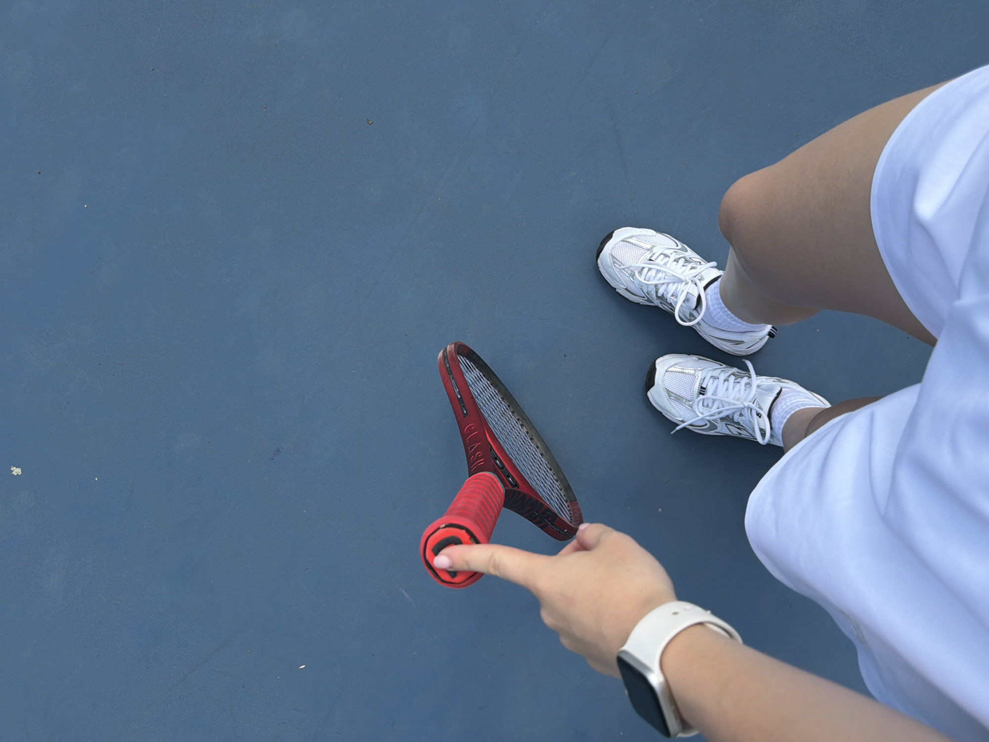 This is a first-person perspective, overhead shot of a person standing on a blue tennis court. The person is wearing a white skirt, white socks, and white tennis shoes. A hand with a white watch on the wrist is extended, holding the red grip of a tennis racket. The tennis racket is red with black accents and has strings. The background is a plain blue tennis court, showing no distinguishing landmarks or geographical features, making it impossible to determine the location as Warsaw, Poland.