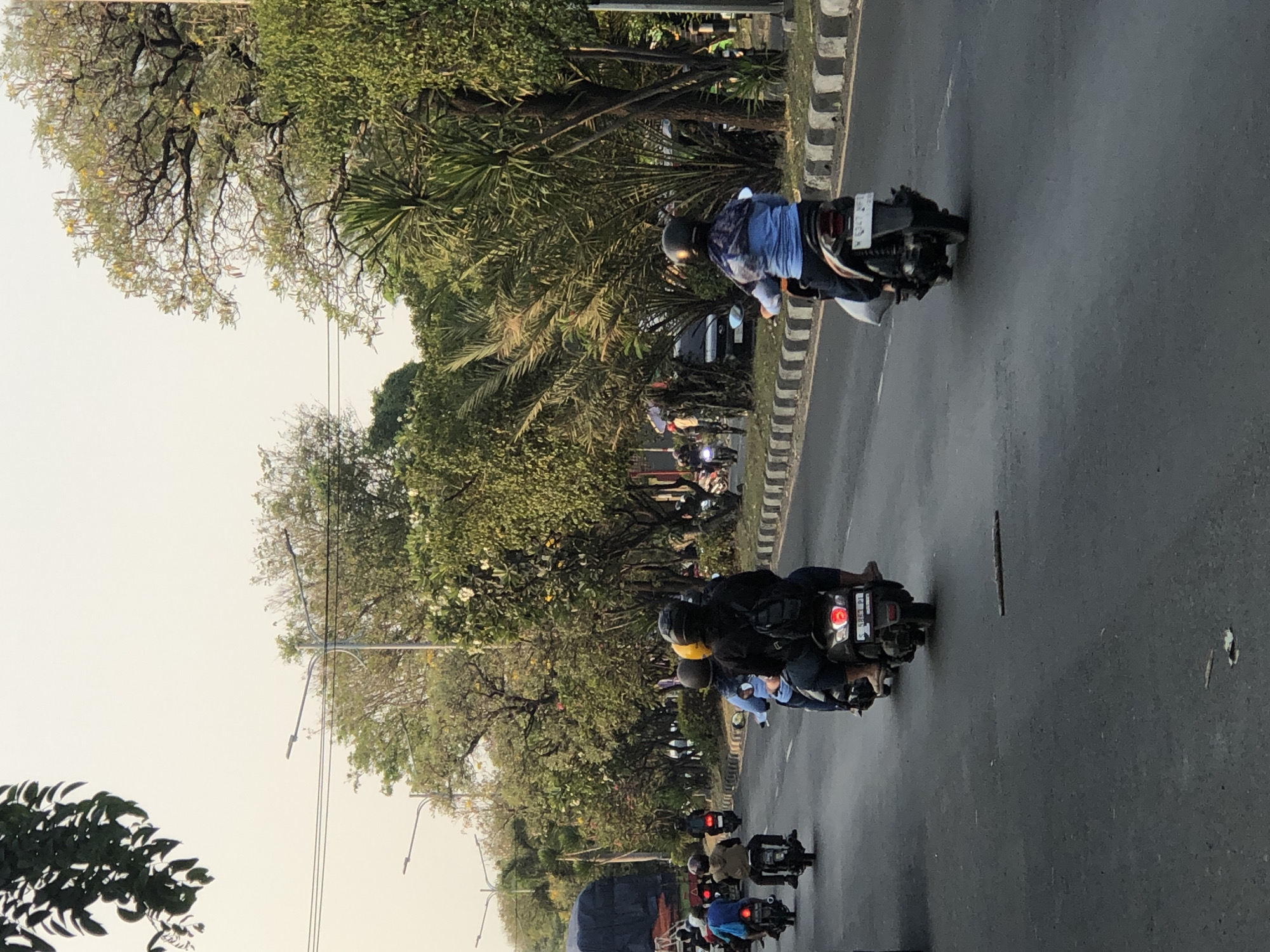 The image depicts a street scene in Ngaban, Indonesia, with multiple motorcycles in motion. The road surface is asphalt, bordered by a gray and white striped curb. Lush green trees and foliage line the right side of the road, with a clear sky overhead.

In the foreground, two individuals are riding on a dark-colored motorcycle. The rider in front wears a black helmet and a black hoodie, with a backpack visible. The passenger behind wears a blue patterned shirt and a dark helmet.

Further ahead on the road, several other motorcycles are visible. To the left, a motorcycle with a rider wearing a blue shirt and helmet is seen. Behind it, another motorcycle with two riders, one in a yellow helmet, is present. A line of motorcycles stretches further into the distance on the left side of the road.

To the right of the road, a dark-colored car is parked or moving slowly. The environment suggests a warm, possibly humid climate, consistent with Indonesia. Streetlights are also visible along the road. The overall impression is a busy street with everyday transportation in a tropical urban setting.