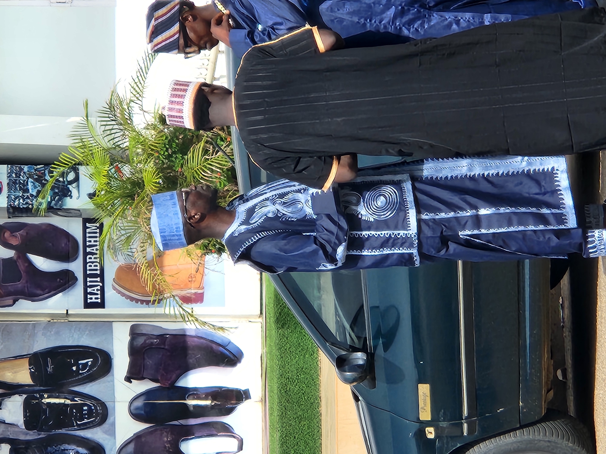 The image shows three men standing next to a dark blue car, seemingly posing for a photo. The men are dressed in traditional African attire. The man closest to the camera wears a blue outfit with intricate white patterns and a blue cap. The second man is in a black robe with brown trim and a white and maroon patterned cap. The third man, further back, is in a blue outfit and a striped cap.
A palm plant stands behind the men, partly obscuring a shop sign displaying various shoes. The sign has the words "HAJI IBRAHIM" on it.
The car's side is visible, showing a "Prestige" emblem. The scene appears to be in an urban setting, although the specific location is not identifiable.