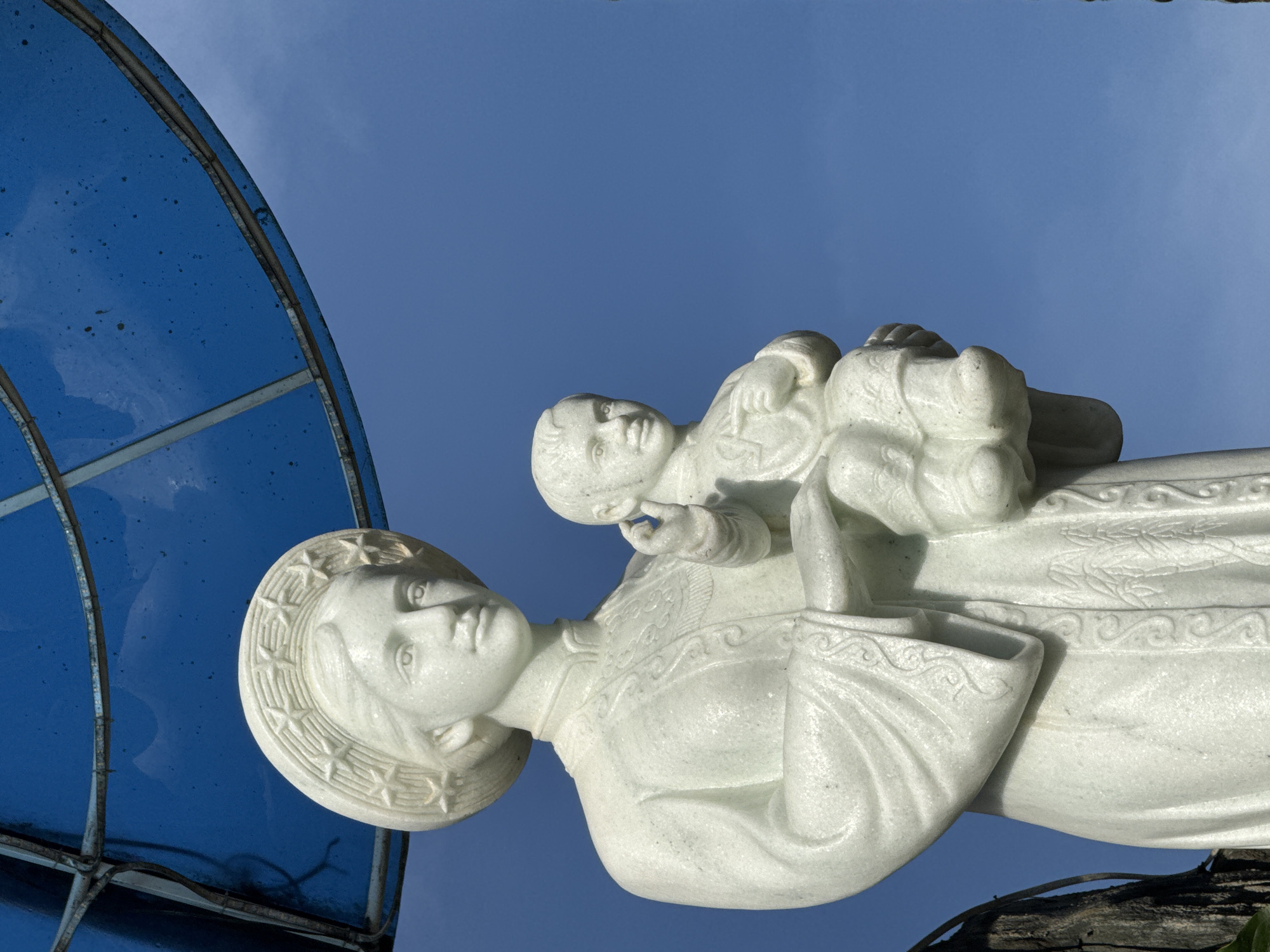 A white marble statue of Mary holding the infant Jesus stands against a bright blue sky. The statue is positioned under a blue, circular canopy supported by metal framing. Mary wears a hat adorned with stars and a traditional Vietnamese dress with intricate embroidered patterns on the collar and sleeves. Jesus is depicted as a young child held in her arms, with his right hand raised in a blessing gesture. The statue exhibits fine detail in the sculpting of the facial features and drapery. The background is a clear, cloudless blue sky, with hints of dark foliage and tree trunks visible on the left edge of the frame. The overall scene appears to be outdoors, likely in a public space or religious site within the city of Đa Thọ, Vietnam.