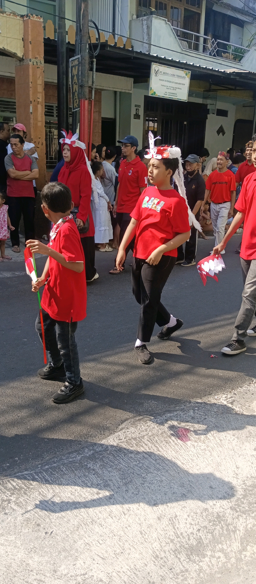 This image depicts a street scene in the city of Malang, Indonesia, likely during a parade or cultural event. Several people are walking on a paved street. In the foreground, a young boy in a red t-shirt and jeans is holding a small Indonesian flag on a green pole. To his right, another person, also in a red t-shirt and black pants, is walking and holding a red and white flag with a zigzag edge. Both individuals appear to be wearing festive headbands with white and red feathers.
In the background, a crowd of people are visible, many of them wearing red clothing, suggesting a coordinated theme. Behind the crowd, buildings with balconies and signage can be seen. One sign, visible on the building, reads "PT. MIONS CAKRA TEKNOLOGI PRODUSER DAN PENYEDIA ALAT DASAR LABORATORIUM". The street is lined with what appear to be utility poles, and shadows from people and objects are cast on the ground, indicating a sunny day. The overall atmosphere suggests a lively community gathering.