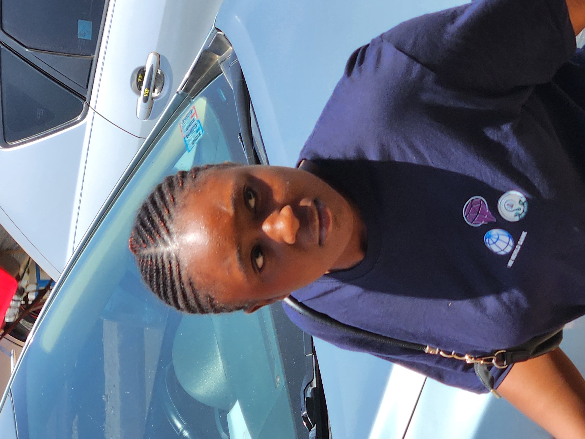 The image depicts a person with dark skin and braided hair standing next to a light-colored car, potentially light blue or silver. The person is wearing a dark blue t-shirt with what appear to be colorful graphic pins or designs on the right side of the chest. A black bag strap is visible across the person's body. The car's windshield is reflecting the sky and potentially other objects in the surrounding area. The location appears to be outdoors, possibly a city street. The exact city is unknown.