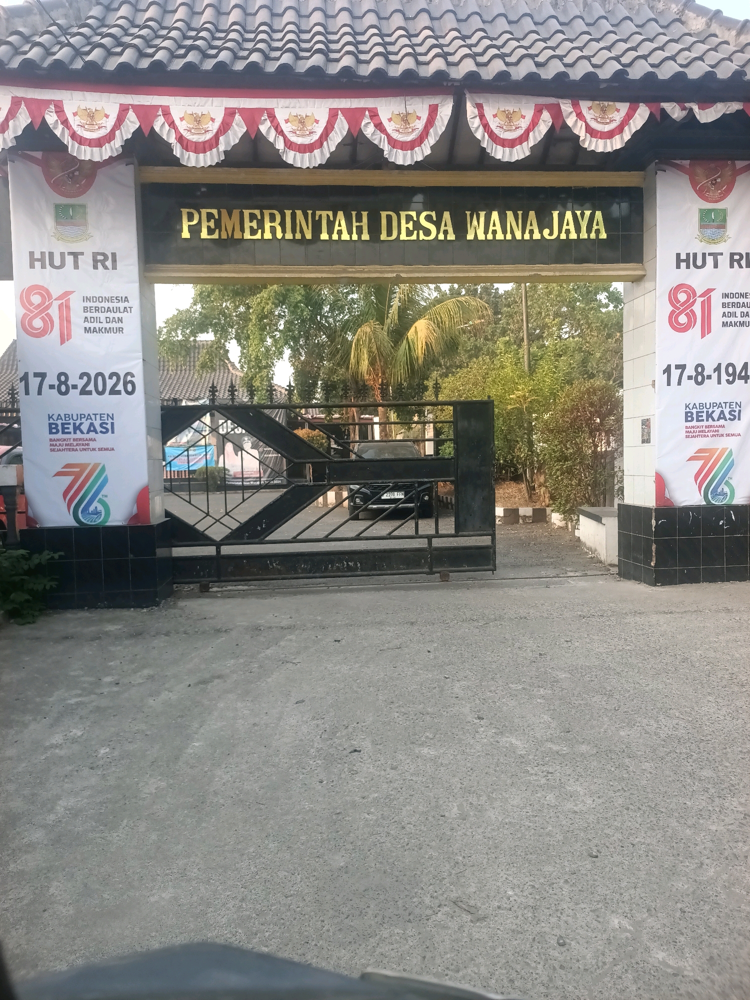 The image shows the entrance to the "PEMERINTAH DESA WANAJAYA" (Wanajaya Village Government) in Wanajaya, Indonesia. Two large banners are prominently displayed on either side of the entrance gate. The banner on the left, a white vertical banner, has the text "HUT RI" at the top, followed by the number "81" in large red font. Below this, it reads "INDONESIA BERDAULAT ADIL DAN MAKMUR" and "17-8-2026", with "KABUPATEN BEKASI" and further text below. The banner on the right is similar, also bearing "HUT RI" and the number "81", with additional text including "INDONESIA BERDAULAT ADIL DAN MAKMUR" and "17-8-194". It also has "KABUPATEN BEKASI" and accompanying text. Both banners feature a stylized number 7 and what appears to be a depiction of the Indonesian archipelago.
The entrance gate is a black metal structure with an intricate geometric pattern. Behind the gate, there is a paved area with trees and vegetation visible in the background. The building above the entrance is roofed with dark tiles and adorned with red and white decorative bunting featuring the Garuda Pancasila emblem. A dark-colored vehicle is partially visible through the gate. The overall scene depicts a government building entrance decorated for a celebration, likely Indonesian Independence Day (HUT RI).