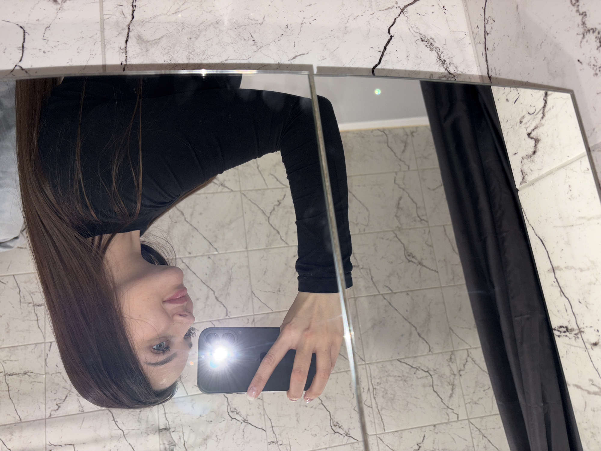 A woman is captured taking a mirror selfie, seen from the chest up and rotated to be sideways. Her long, dark brown hair falls over her shoulders. She wears a plain black long-sleeved top. Her face, featuring blue eyes, light pink lips, and long eyelashes, is visible as she looks towards her phone. She holds a dark-colored smartphone in her right hand, with its bright flash illuminated, positioned in front of her.
The reflection is cast in a mirror that appears to be composed of two vertical panels, indicated by a faint seam in the middle. The background reflected in the mirror consists of walls covered in white marble-patterned tiles with distinct dark grey and black veins. To the right side of the mirror, a dark, possibly black, shower curtain or similar fabric is visible. The scene suggests an indoor setting, likely a bathroom or dressing area, illuminated by artificial light, primarily from the phone's flash. No visible text is present in the image.