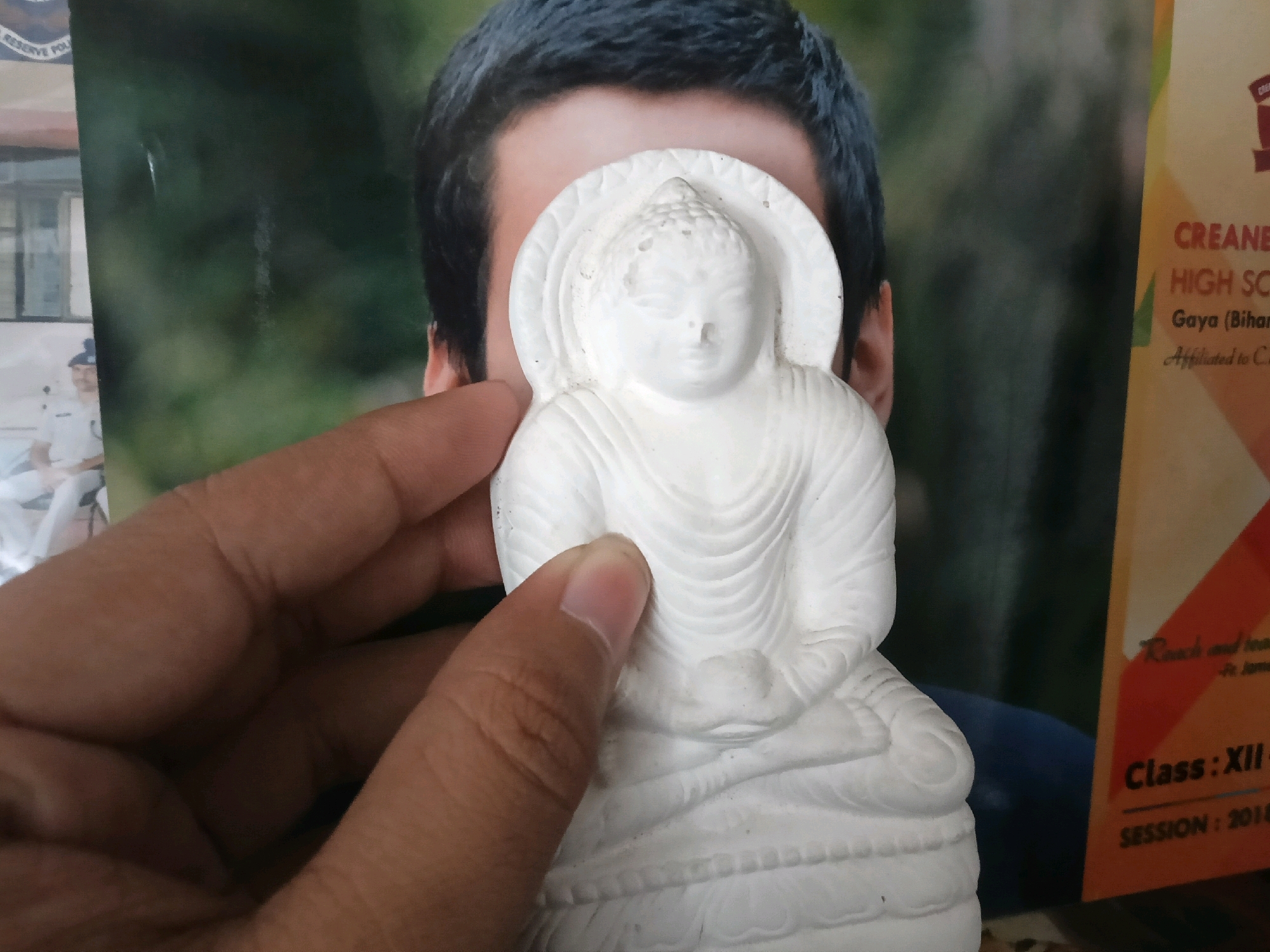 A close-up, slightly angled shot shows a hand holding a small, white statue of Buddha in front of a blurred background. The statue is positioned to obscure the face of a person in the background, whose dark hair and eyes are visible. To the right of the statue, a portion of a yellow and orange poster with text is visible. The text on the poster includes "CREAN... HIGH SC...", "Gaya (Bihar...", "Reach and le...", "Class: XII...", and "SESSION : 201...". The scene appears to be indoors, with the lighting coming from the front. The overall impression is that of a casual, informal photograph. The location of Manpur, India is mentioned in the prompt, but there is no visual information in the image to confirm or deny this specific location.