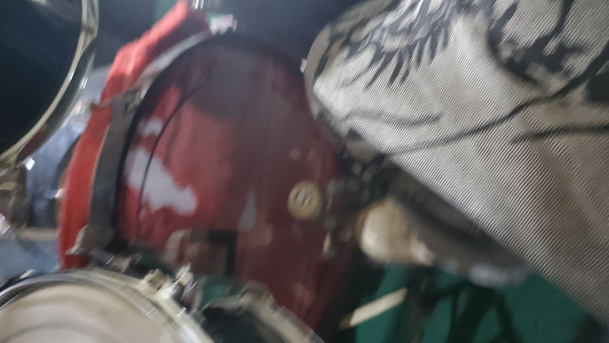 The image depicts a close-up view of a drum set. In the foreground, a section of a drum with a red and white finish is visible, along with a portion of a drumstick or similar object. The background is dark and out of focus, with hints of metallic elements that could be part of the drum hardware or other instruments. The overall lighting is dim, suggesting an indoor setting, possibly a music studio or a performance space. The scene does not contain any elements that would indicate a specific location in Jalingo, Nigeria.