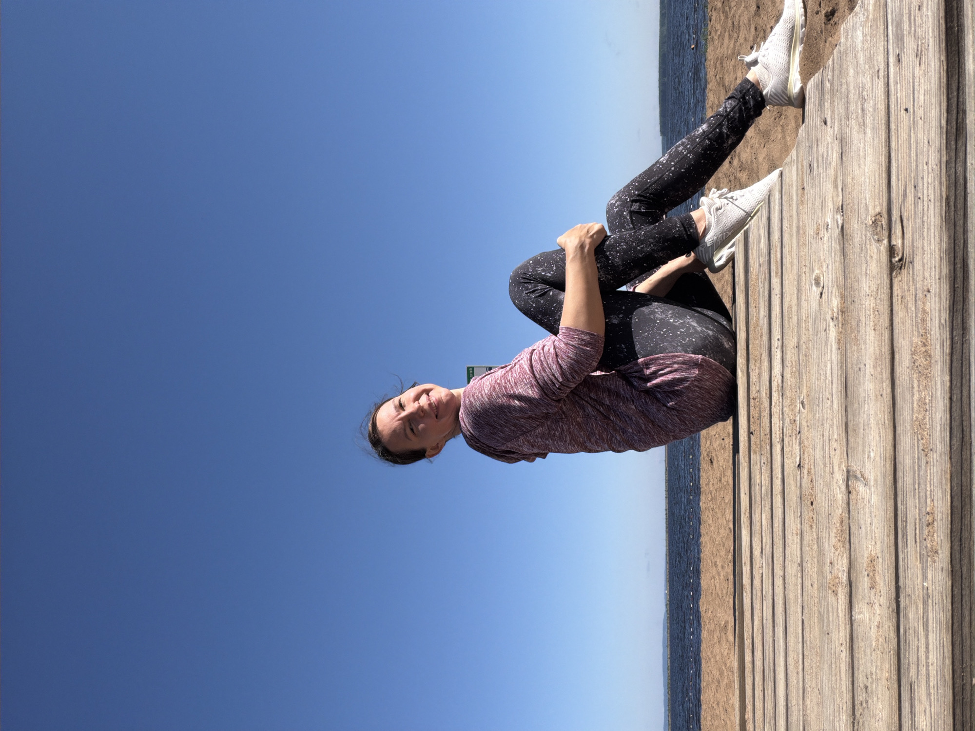 A woman is sitting on a wooden boardwalk near the beach, with a clear blue sky above. She is wearing a pink and white hooded sweatshirt and black leggings speckled with white. Her legs are crossed, and she is resting her hands on her knees. She is looking over her shoulder at the camera with a smile. The boardwalk is made of weathered wooden planks and is situated on a sandy beach. In the background, there is a body of water, likely the Kama River, and a distant shoreline. The overall scene suggests a casual outdoor activity in a coastal area. Based on the context of Chaikovsky, Russia, this location could be along the Kama River, possibly in a recreational area or park.