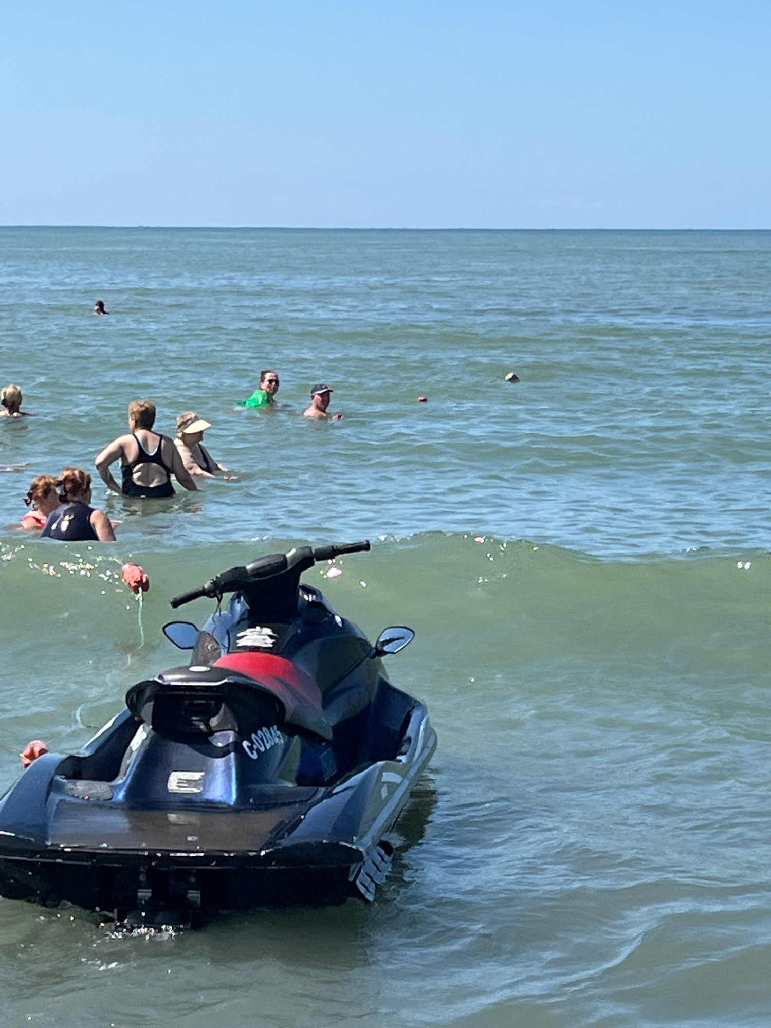 A dark blue and red jet ski is partially submerged in the water in the foreground of the image. The jet ski has a license plate that reads "C-02945". In the middle ground, several people are in the ocean, some swimming and others standing in the water. The water is choppy with small waves. The background consists of a clear blue sky and the horizon where the sky meets the sea. The scene is set at the beach in Ureki, Georgia, known for its magnetic black sand.