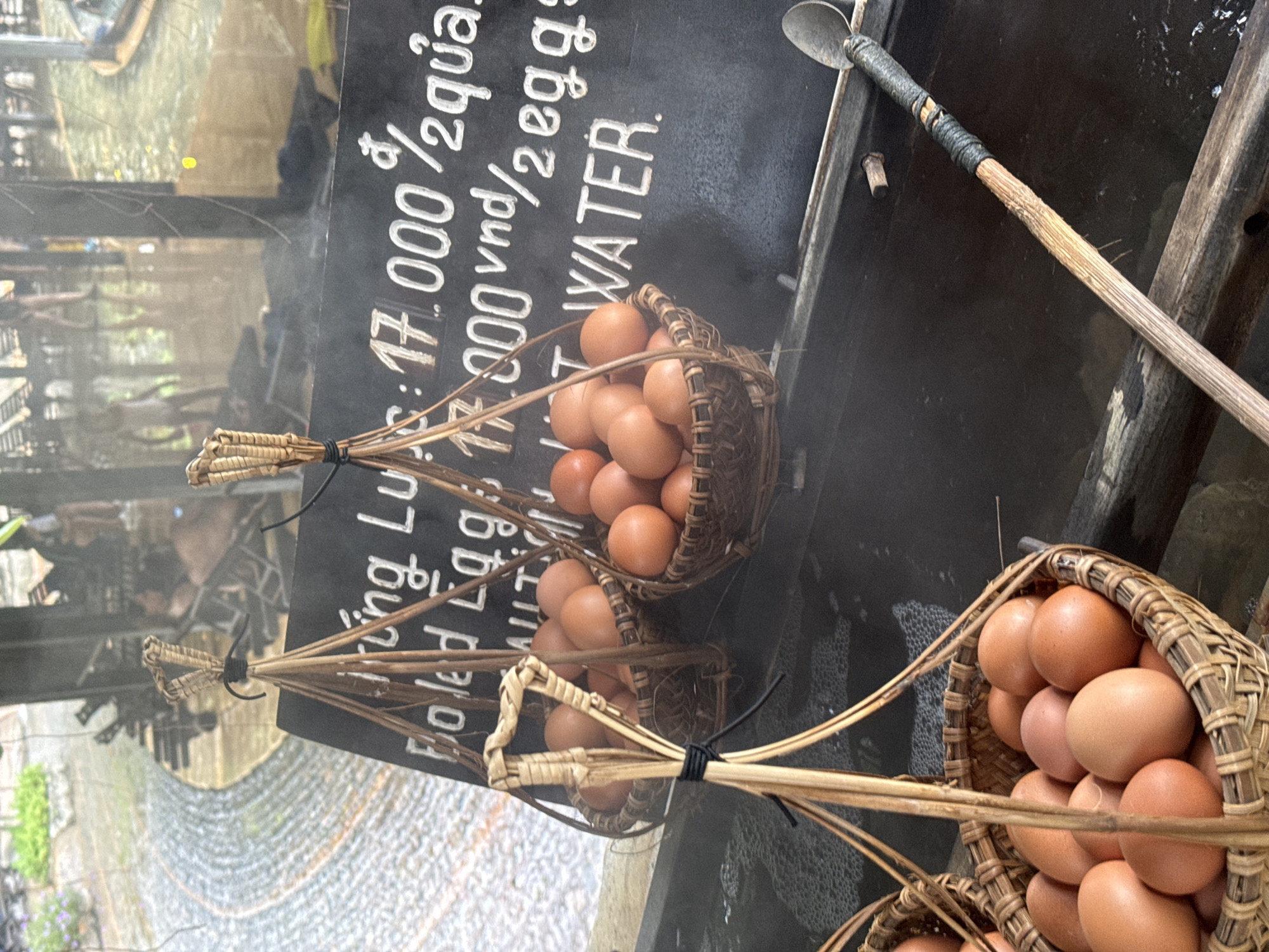 This image depicts an outdoor scene, likely at a market or food stall in Nha Trang, Vietnam. The foreground features two woven bamboo baskets filled with brown eggs, suspended from wooden poles. A dark sign with white lettering is visible behind the baskets. The sign appears to be an advertisement or price list, with some text legible including "Boiled Egg" and "WATER". The background shows more people in a blurry, possibly rainy, environment, with a cobblestone-like pathway and water features. There is steam or mist rising from the area, suggesting a cooking or hot spring environment. The overall impression is one of a traditional or rustic food service setting.