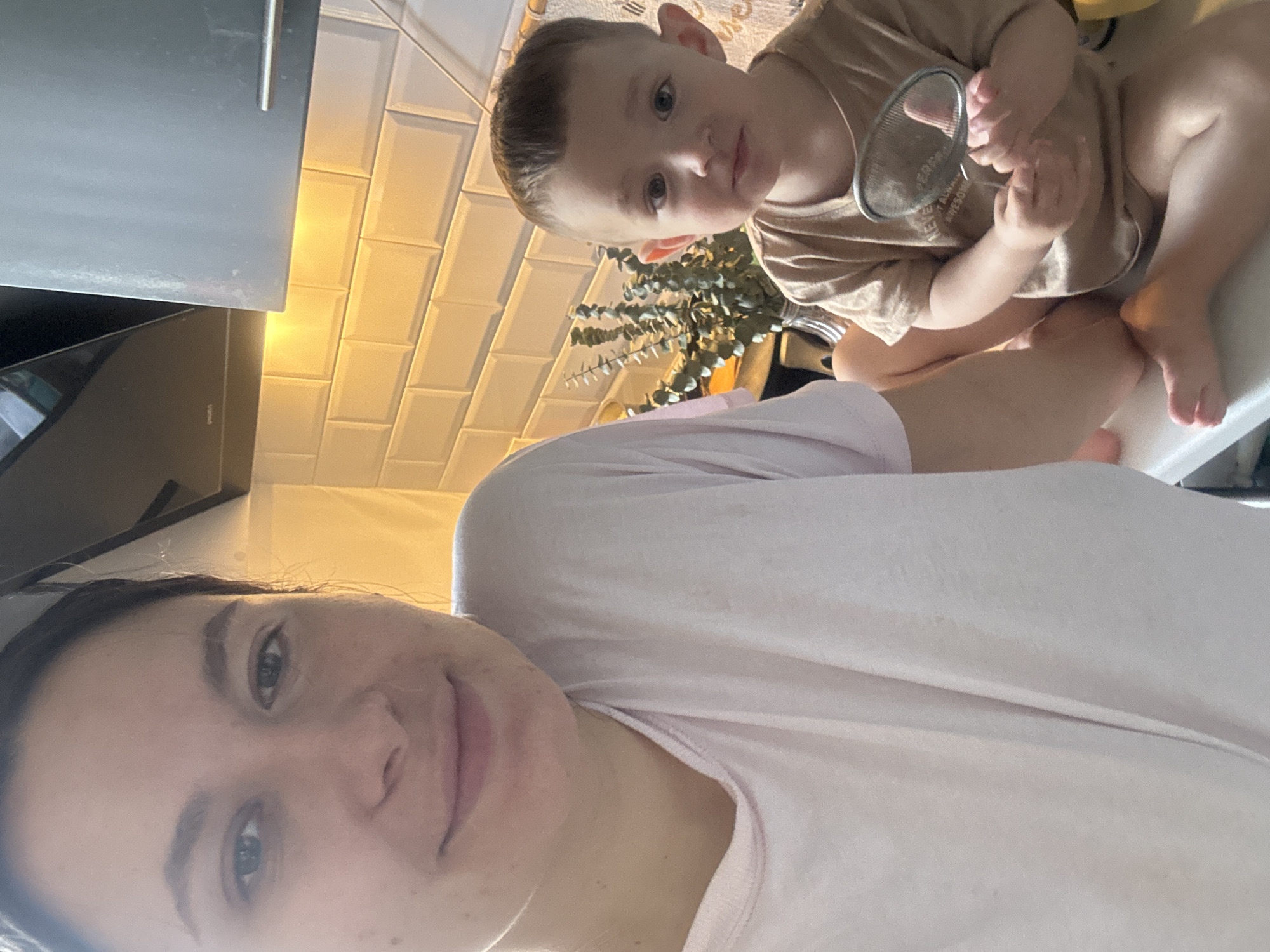 The image shows a woman taking a selfie with a young child. The woman is wearing a light pink shirt. The child is sitting on a countertop, holding a small strainer. The kitchen has white subway tiles on the wall and gray kitchen cabinets. There is greenery in the background. The photo appears to have been taken indoors. There is no information to determine if this image was taken in Odintsovo, Russia.