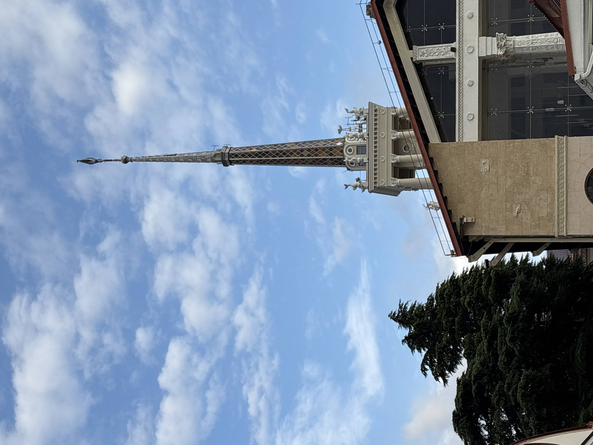 The image shows the upper portion of the Sochi Sea Port building in Sochi, Russia, under a cloudy blue sky. The dominant feature is a tall, ornate spire topping the main tower. The spire is tapering and decorated with a diamond pattern in shades of brown and white. Several small, decorative sculptures resembling figures and dolphins adorn the base of the spire. Below the spire, the building features classical architectural elements, including columns and decorative friezes. To the left of the main tower, a large, dark green evergreen tree is visible, its branches dense and full. The building itself has a textured, light brown stone facade on the left, with large, dark windows on the right section, framed by white decorative elements and columns. The roofline is visible, with red tiles and some metal railings. The overall scene suggests a sunny day with scattered clouds, capturing a prominent landmark in Sochi.