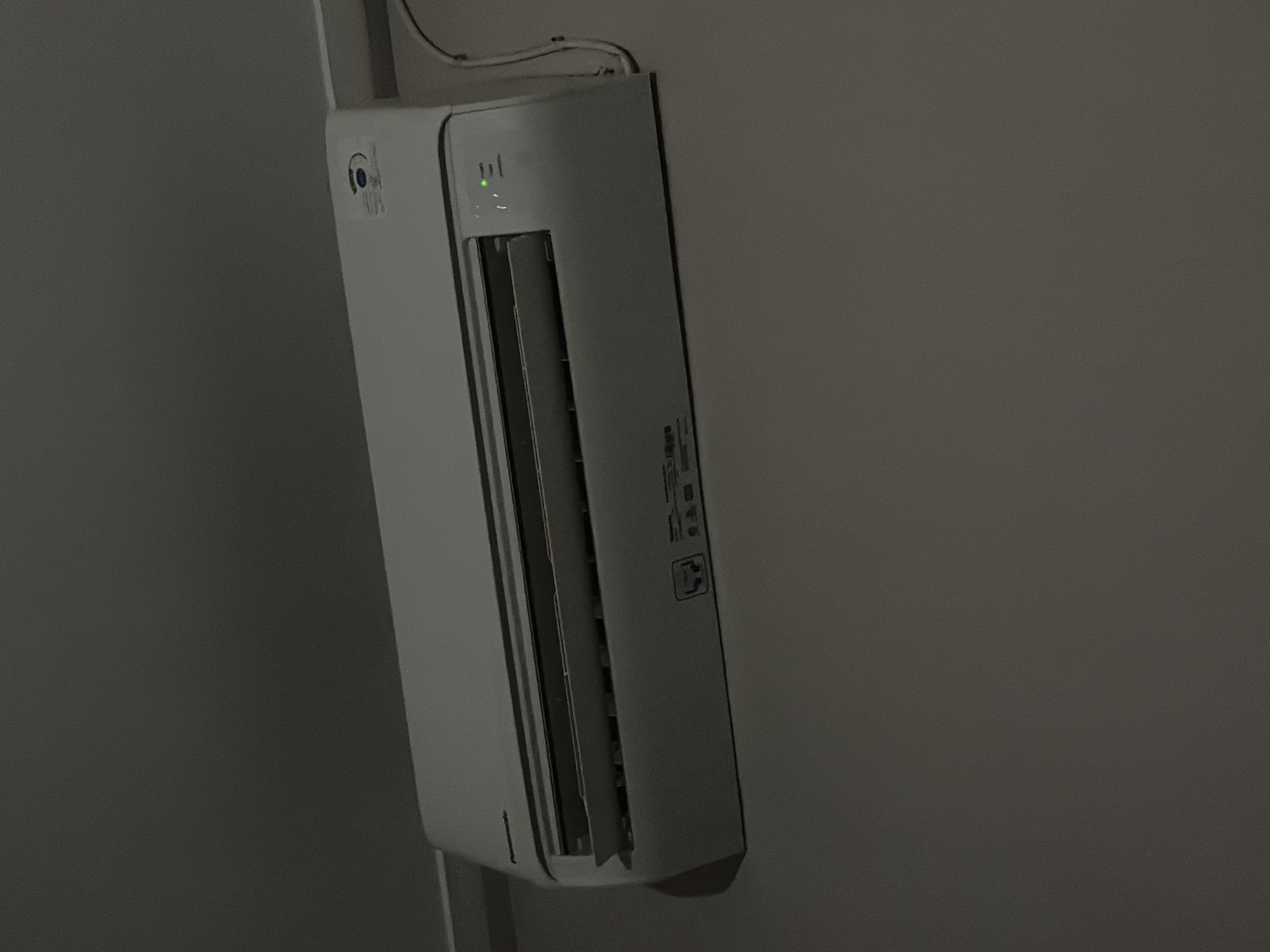 A white Panasonic air conditioner is mounted on a light gray wall, likely in a residential or office setting. The air conditioner has a light gray front panel with horizontal louvers that are slightly open, suggesting it is either in operation or recently powered off. There are indicator lights on the right side of the unit, with a green light illuminated, possibly indicating power or operation. A cable is connected to the right side of the air conditioner and runs down the wall. The wall appears to be painted in a matte finish. The lighting in the room is dim, creating shadows and highlighting the textures of the wall and the air conditioner. There are no people visible in the image. The scene does not provide any specific information about its location within Tupah, Indonesia, beyond being an indoor environment.